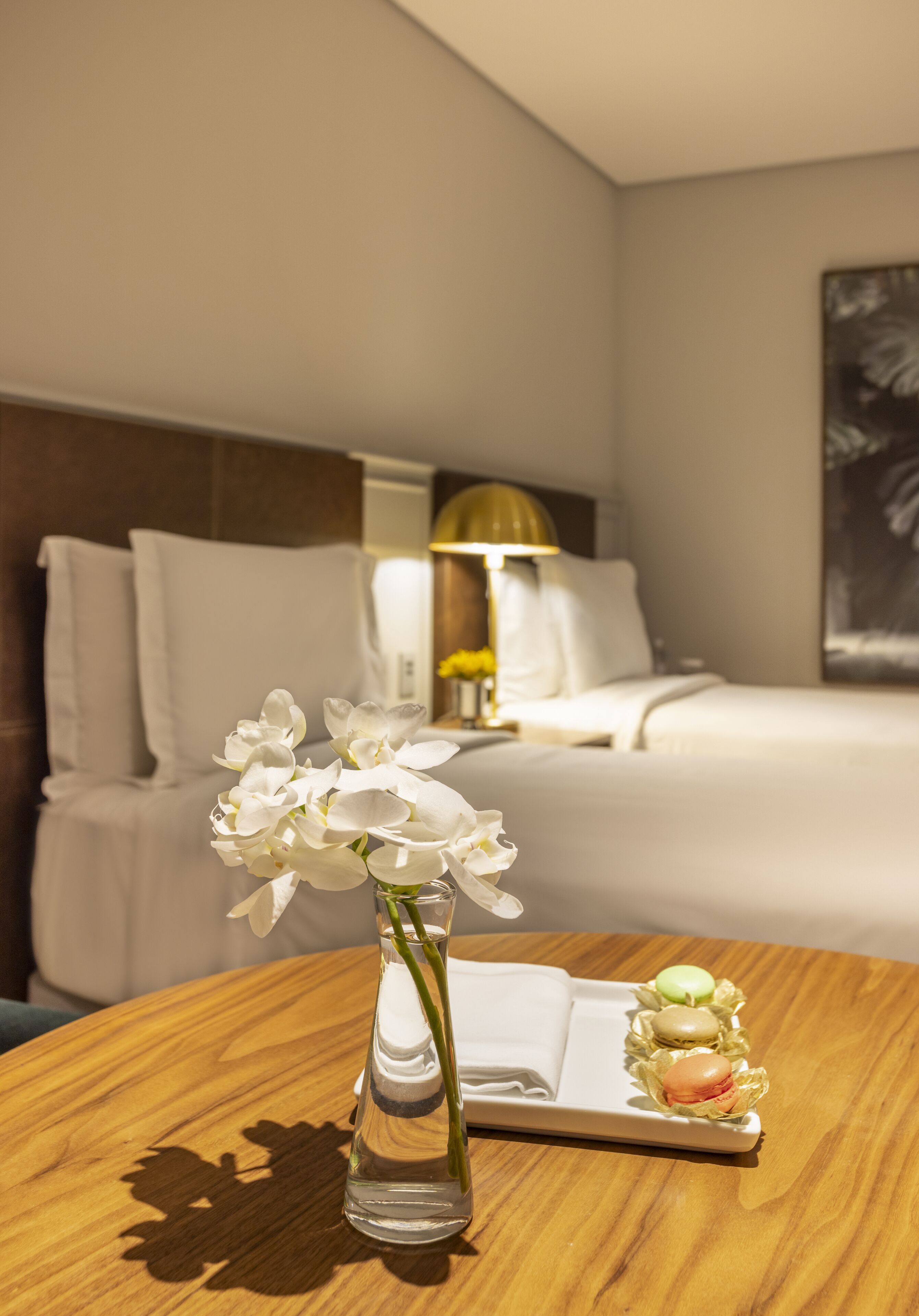 Une chambre d’hôtel avec un lit, une table de chevet avec une lampe et un vase rempli de fleurs, posé sur une table en bois.