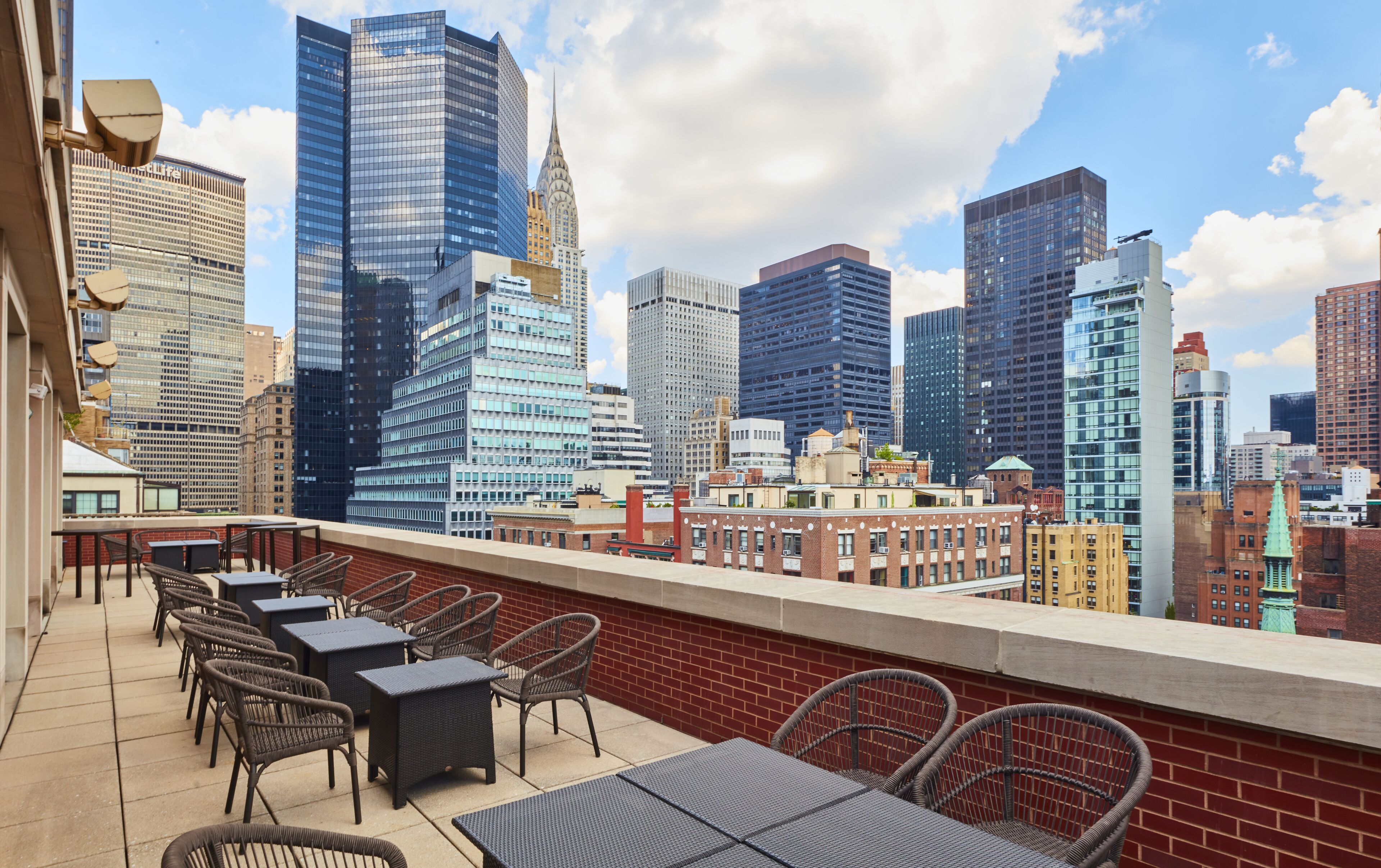 A rooftop terrace with wicker chairs and tables, overlooking a city skyline with modern and historic buildings.