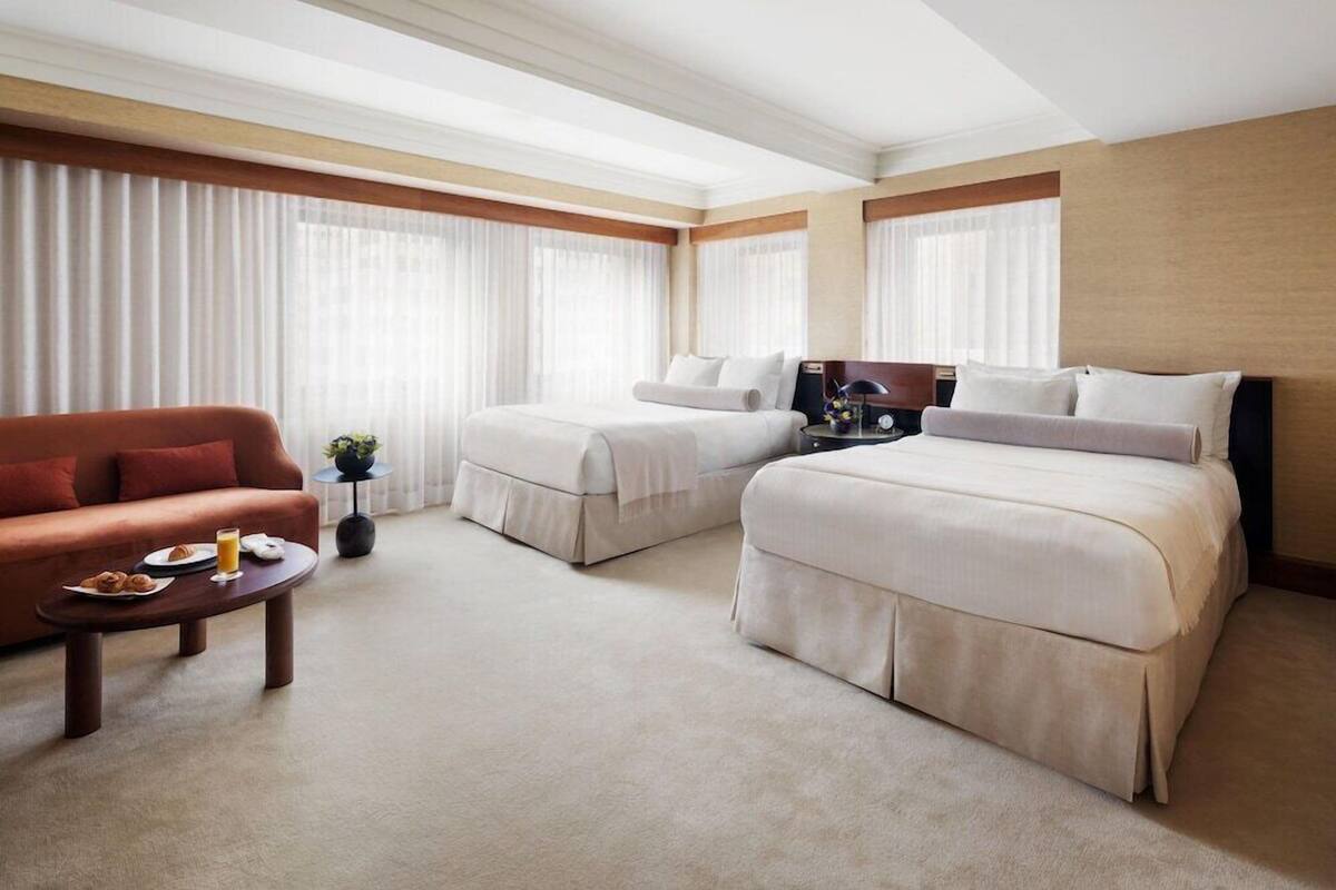 A hotel room with two beds, a sofa, a small table with breakfast, and large windows with sheer curtains.