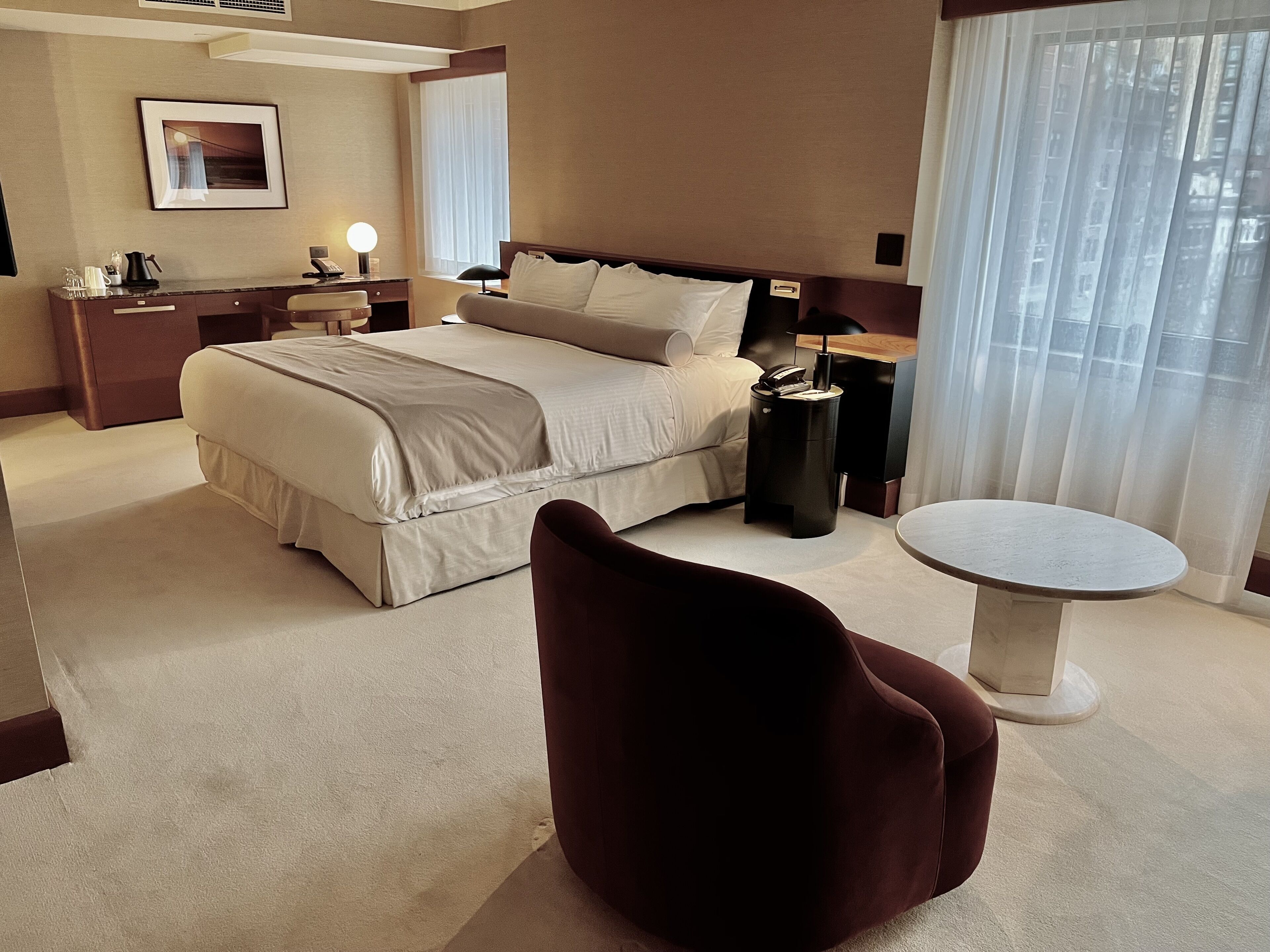 Een hotelkamer met een bed, nachtkastje, lamp, stoel en een klein rond tafeltje.