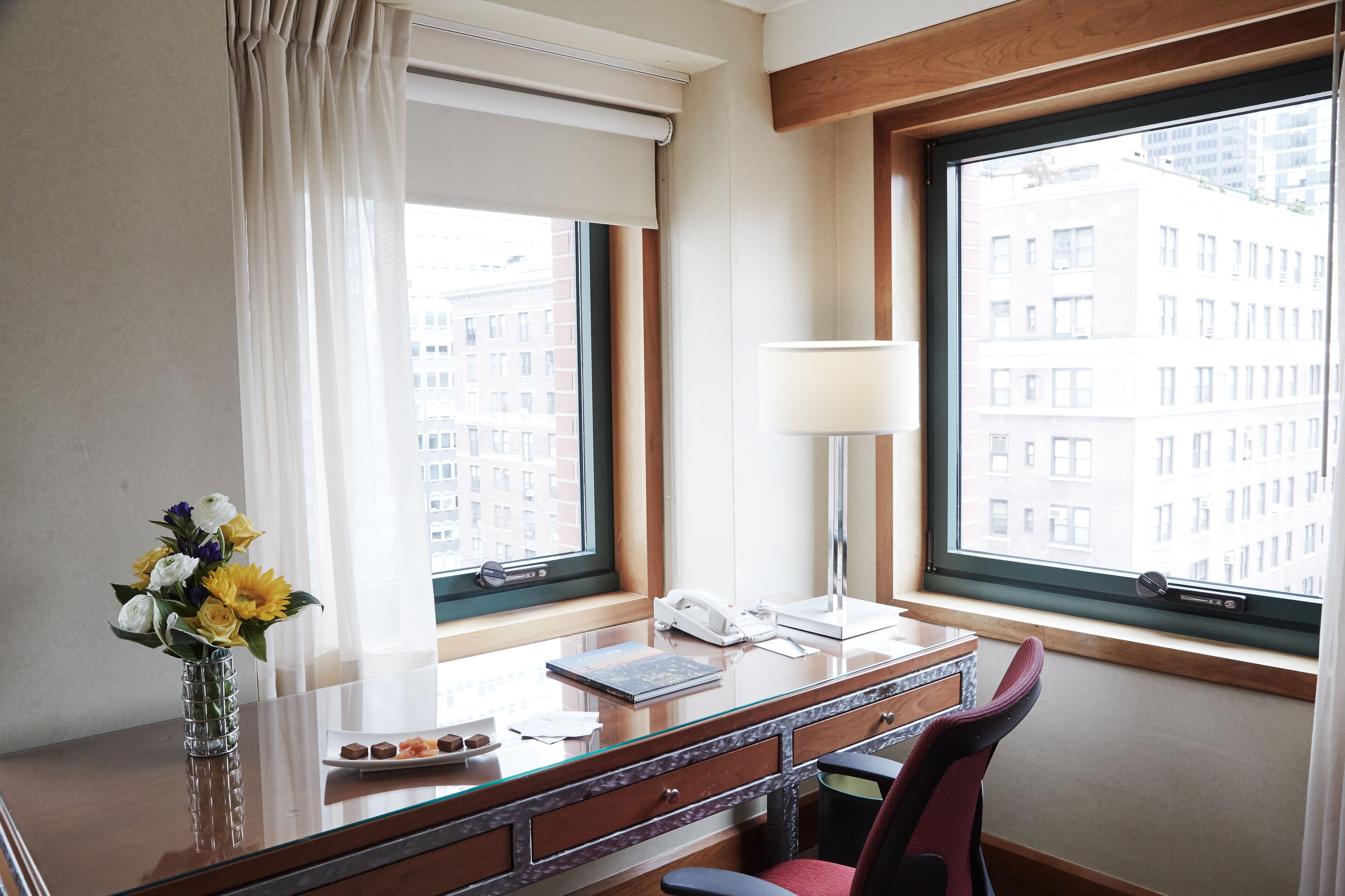 Une chambre d’hôtel équipée d’un bureau, d’un vase de fleurs, d’une lampe et offrant une vue sur les bâtiments à travers de grandes fenêtres.