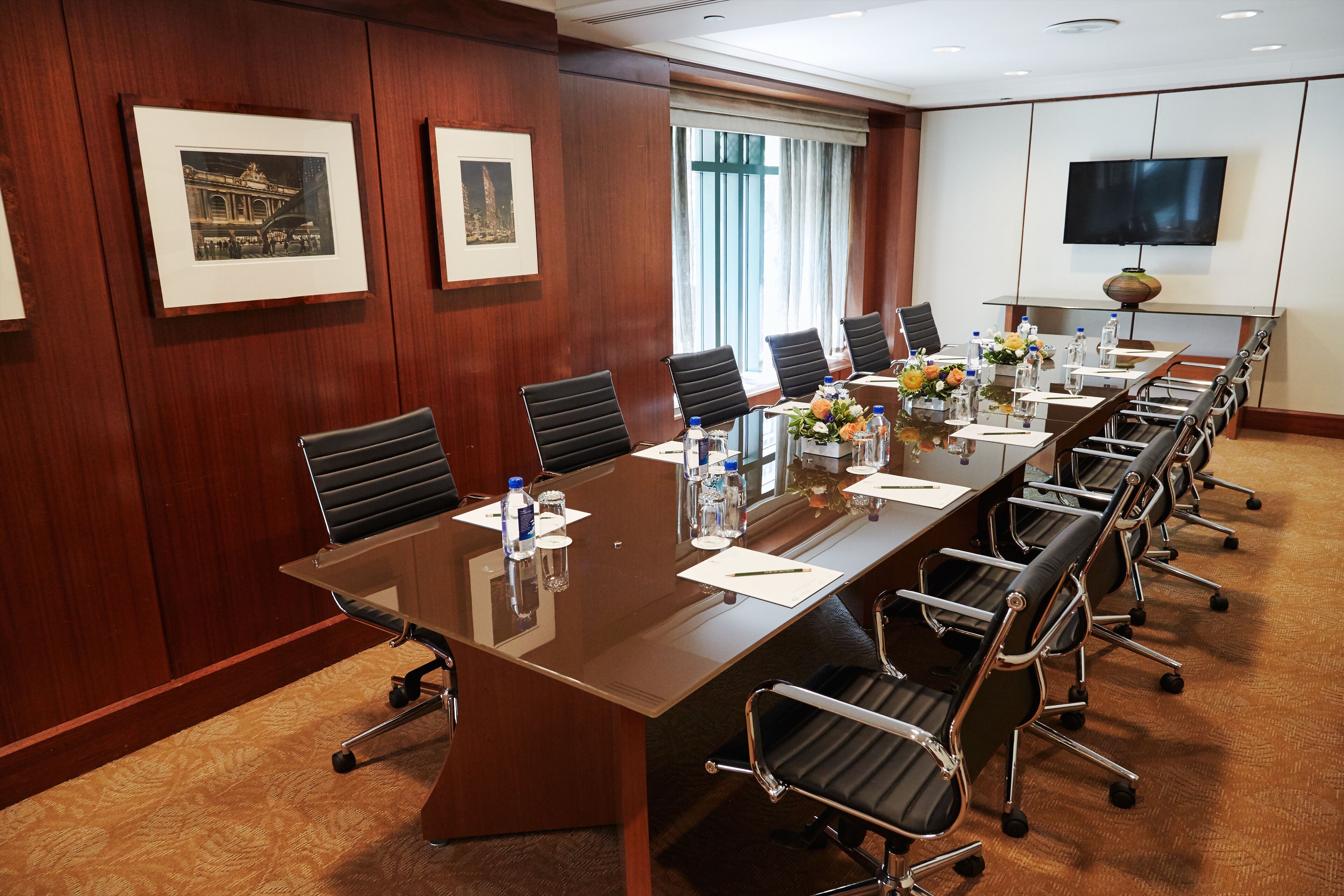 A conference room with a long table, chairs, framed pictures, a TV, and a window with curtains.