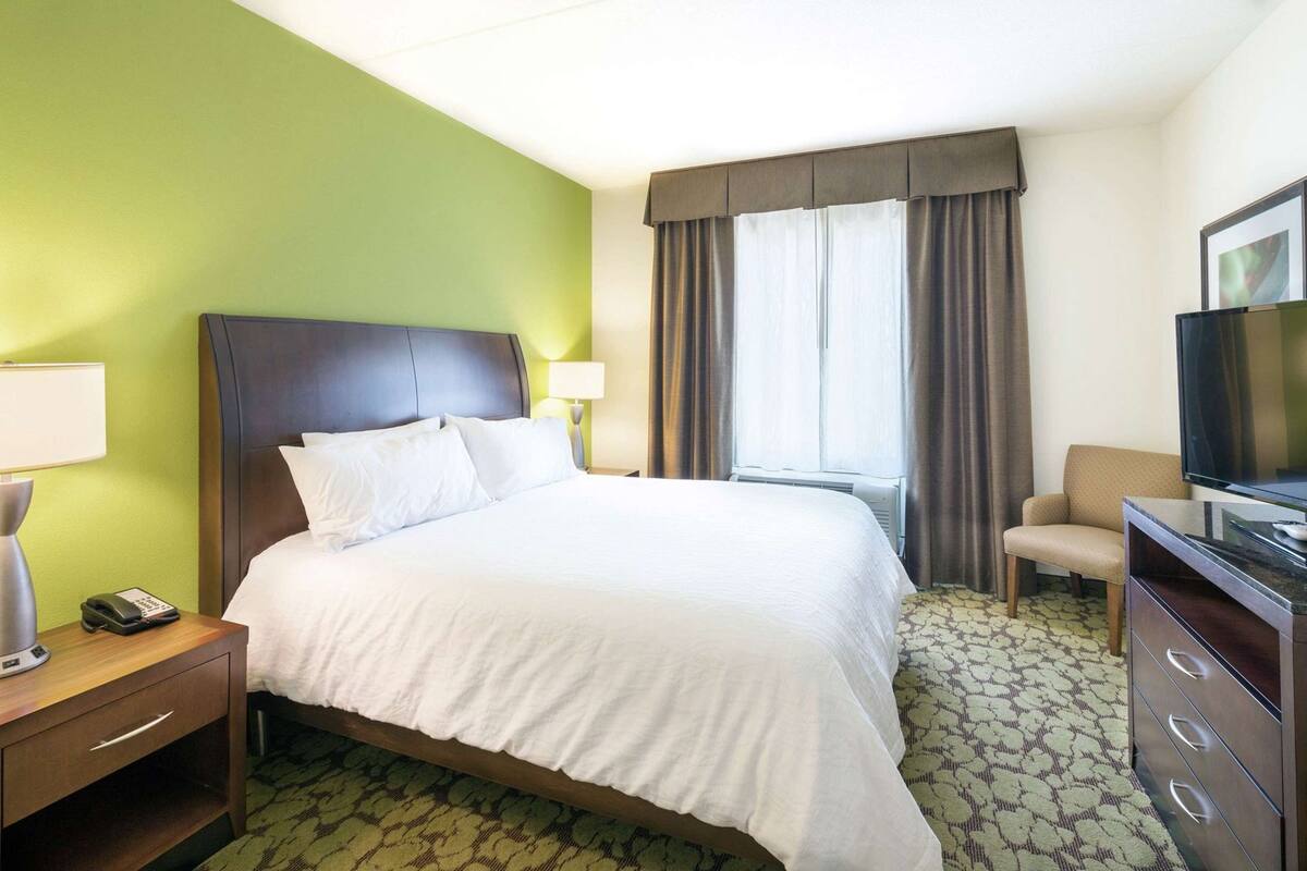 A hotel room with a bed, a desk with a lamp, a chair, a television, and a window with curtains.