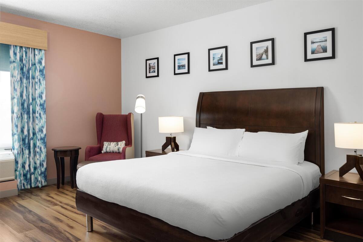 A hotel room with a large bed, a red armchair, a small table, and a lamp. There are four framed pictures on the wall.