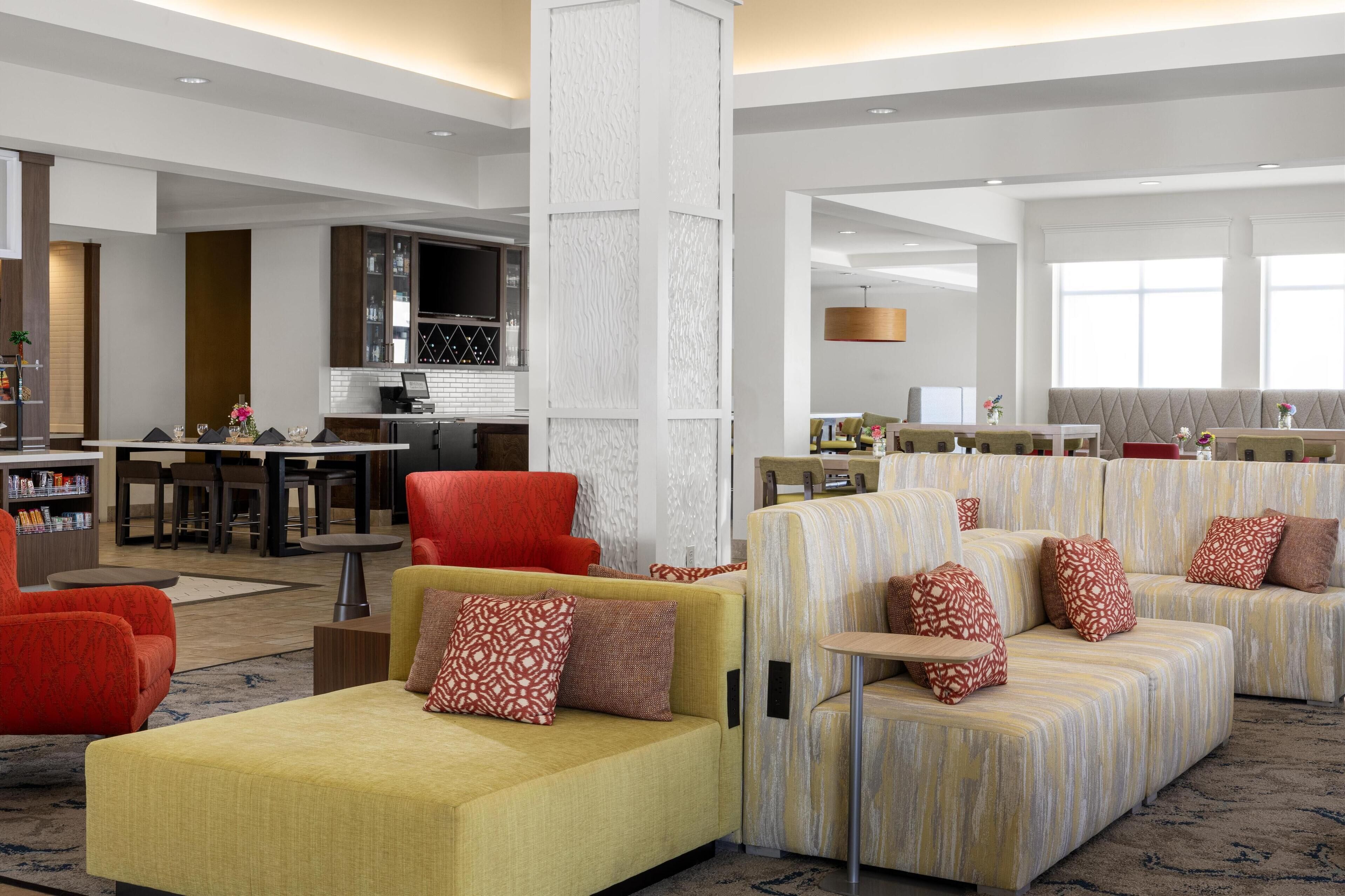 A modern hotel lobby with sofas, armchairs, a dining area, and a kitchen.
