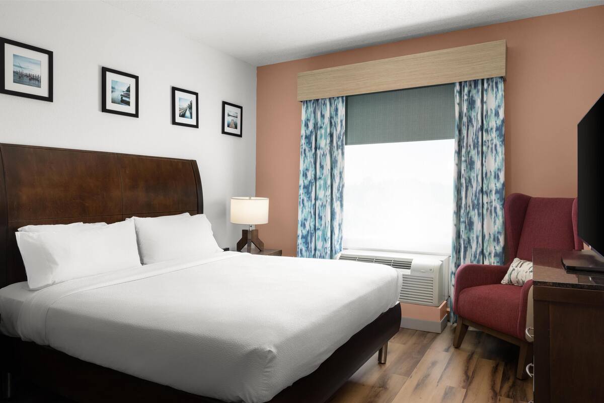 A hotel room with a large bed, a desk, a chair, a window with curtains, and framed pictures on the wall.