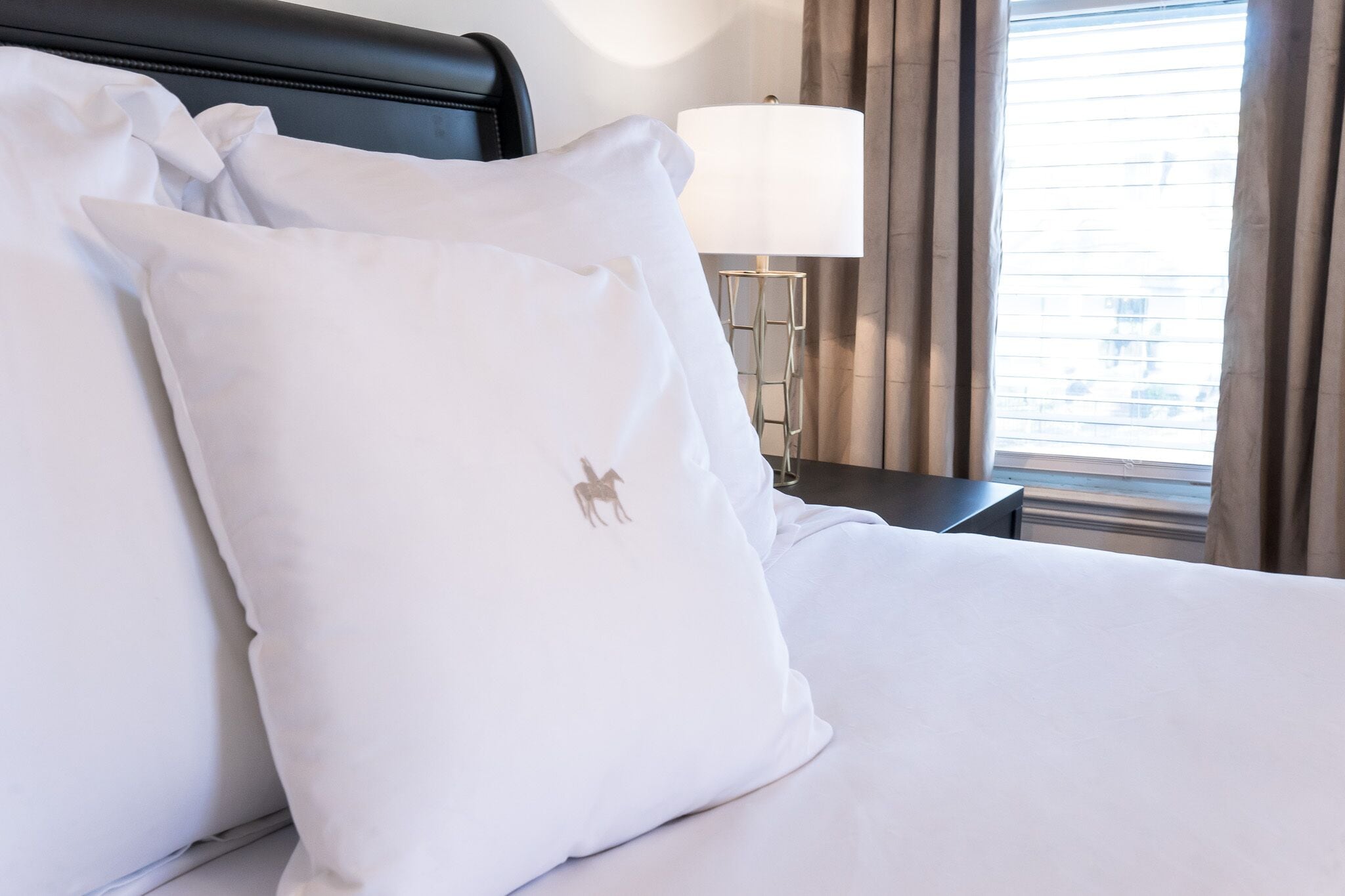A neatly made bed with white linens and a pillow featuring a horse logo.