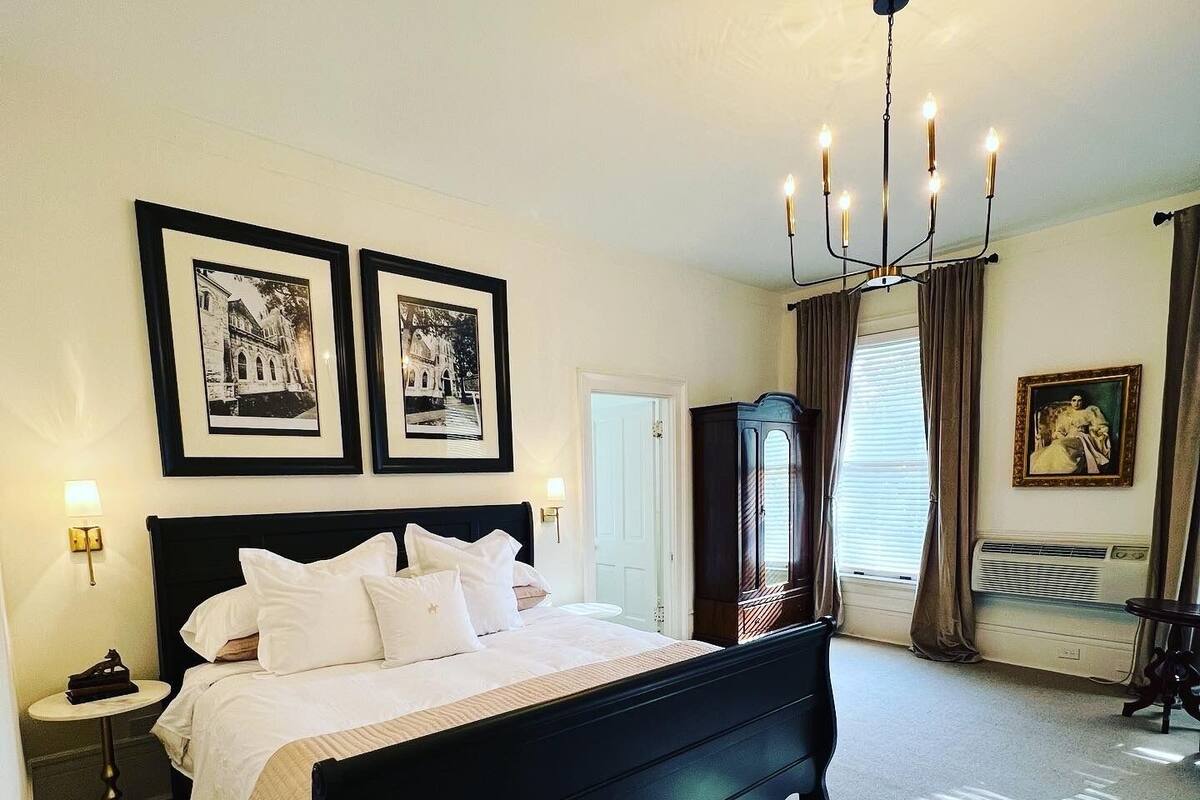 A bedroom with a black bed, two framed pictures on the wall, a chandelier, and a large window with curtains.