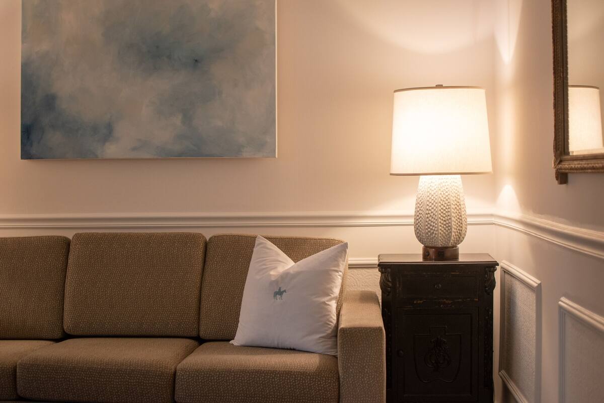 A brown sofa with a white pillow, a textured lamp, and a wall-mounted painting.