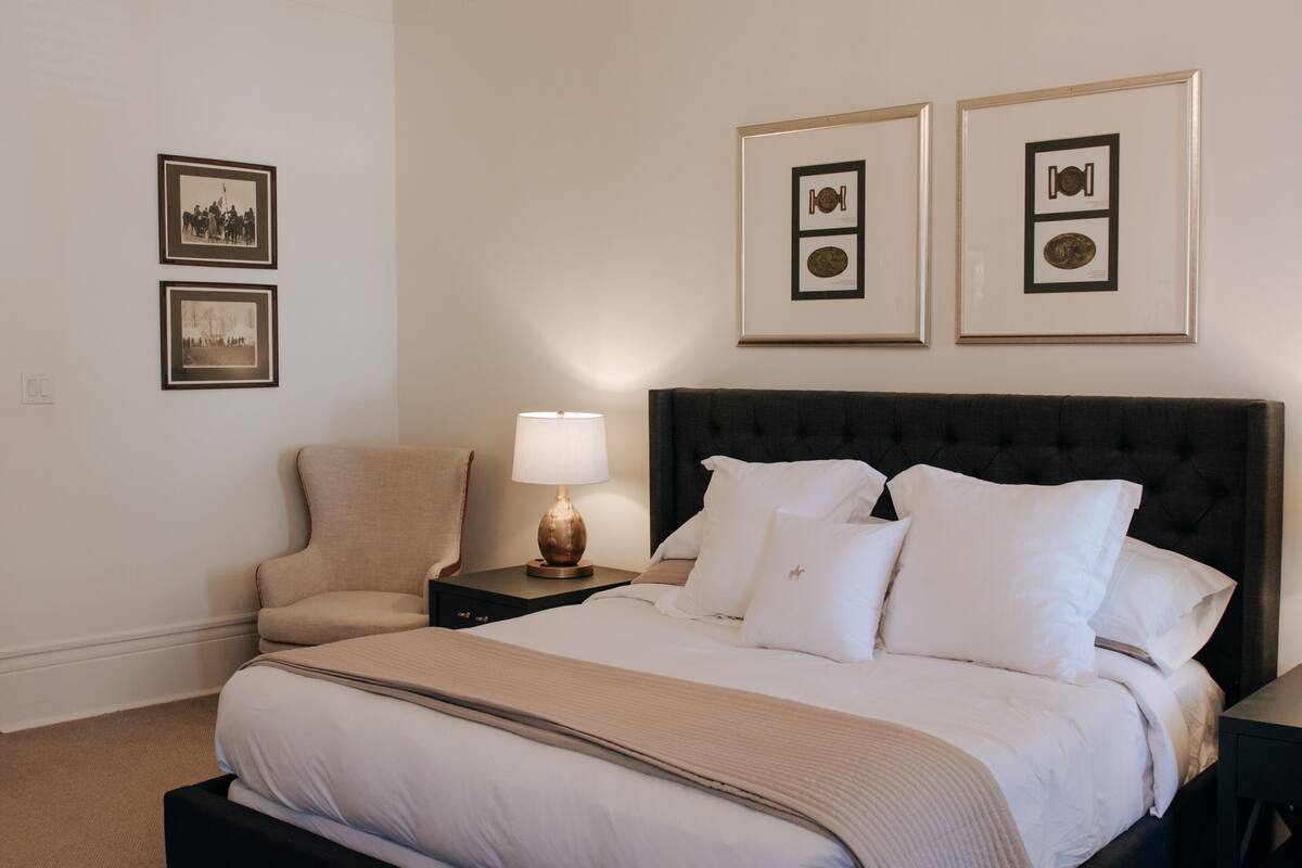 A bedroom with a large bed, a nightstand with a lamp, a chair, and framed pictures on the wall.