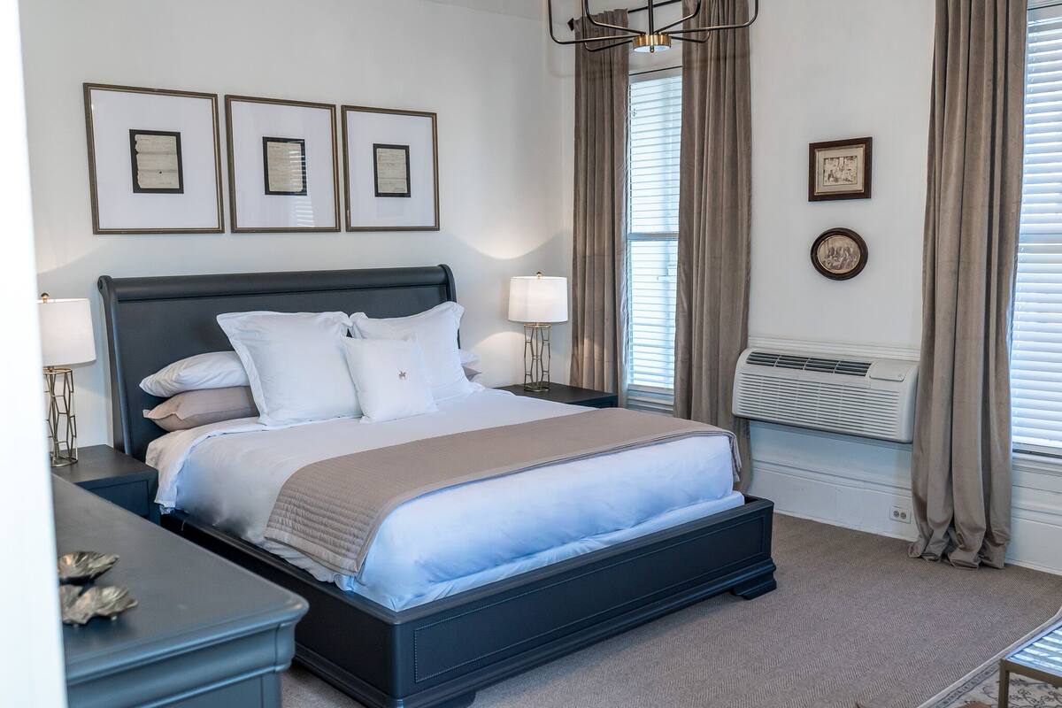 A neatly arranged bedroom with a large bed, bedside lamps, framed artwork on the wall, and a window with curtains.