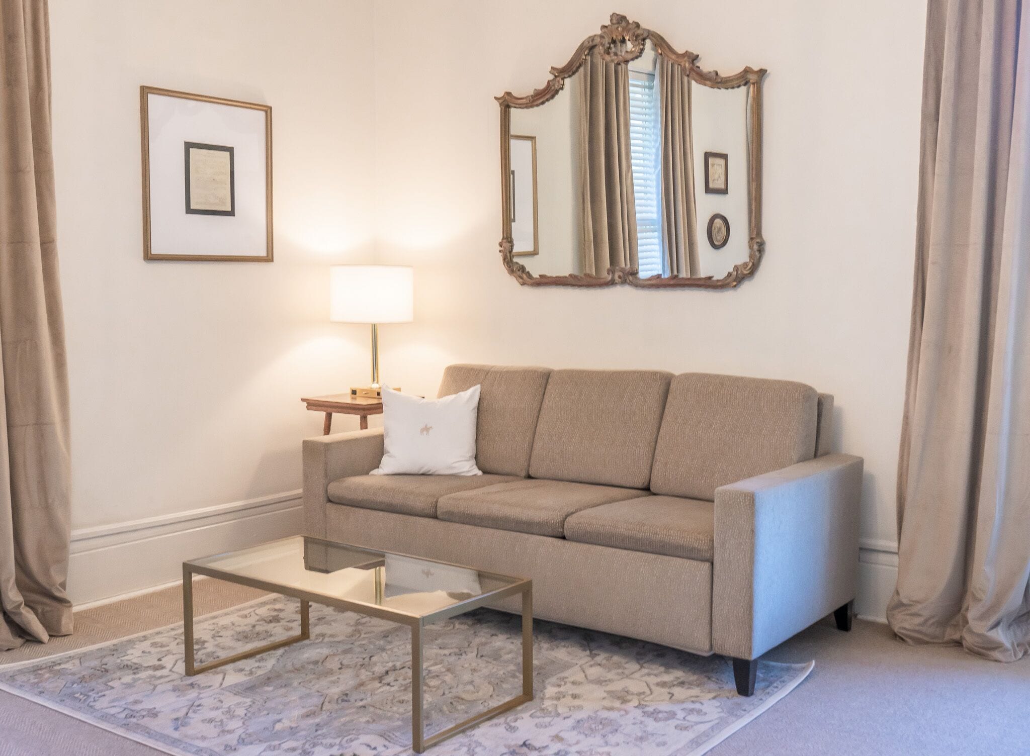 Un salon avec un canapé beige, une table basse en verre, une table d’appoint avec une lampe et un grand miroir au mur.