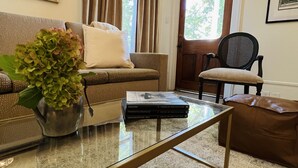 Una sala de estar con un sofá, una silla, una mesa de centro con libros, un jarrón con flores y una puerta con cortinas.