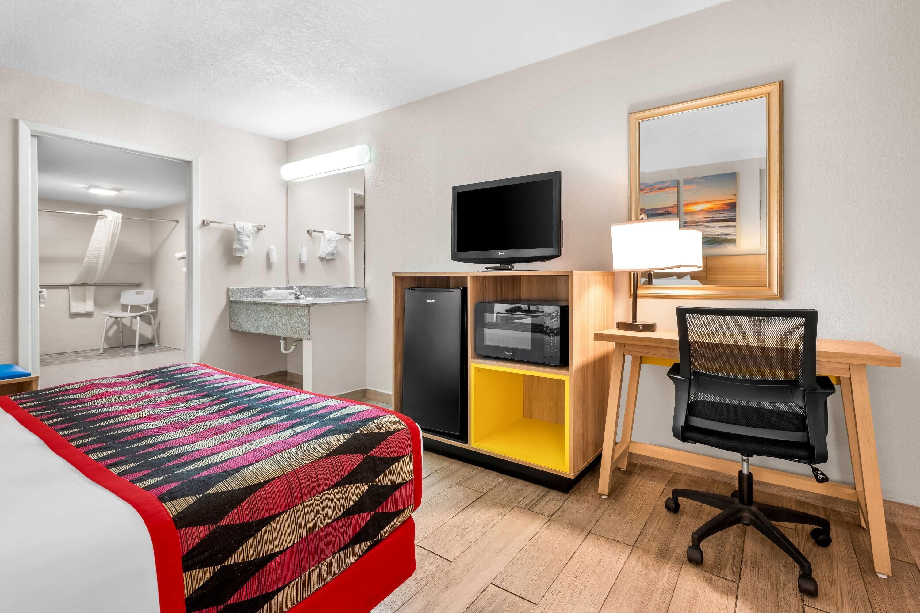 Ein Hotelzimmer mit einem Bett, einem Schreibtisch mit Computer, einem Fernseher, einem Spiegel und einem sichtbaren Badezimmer durch eine offene Tür.