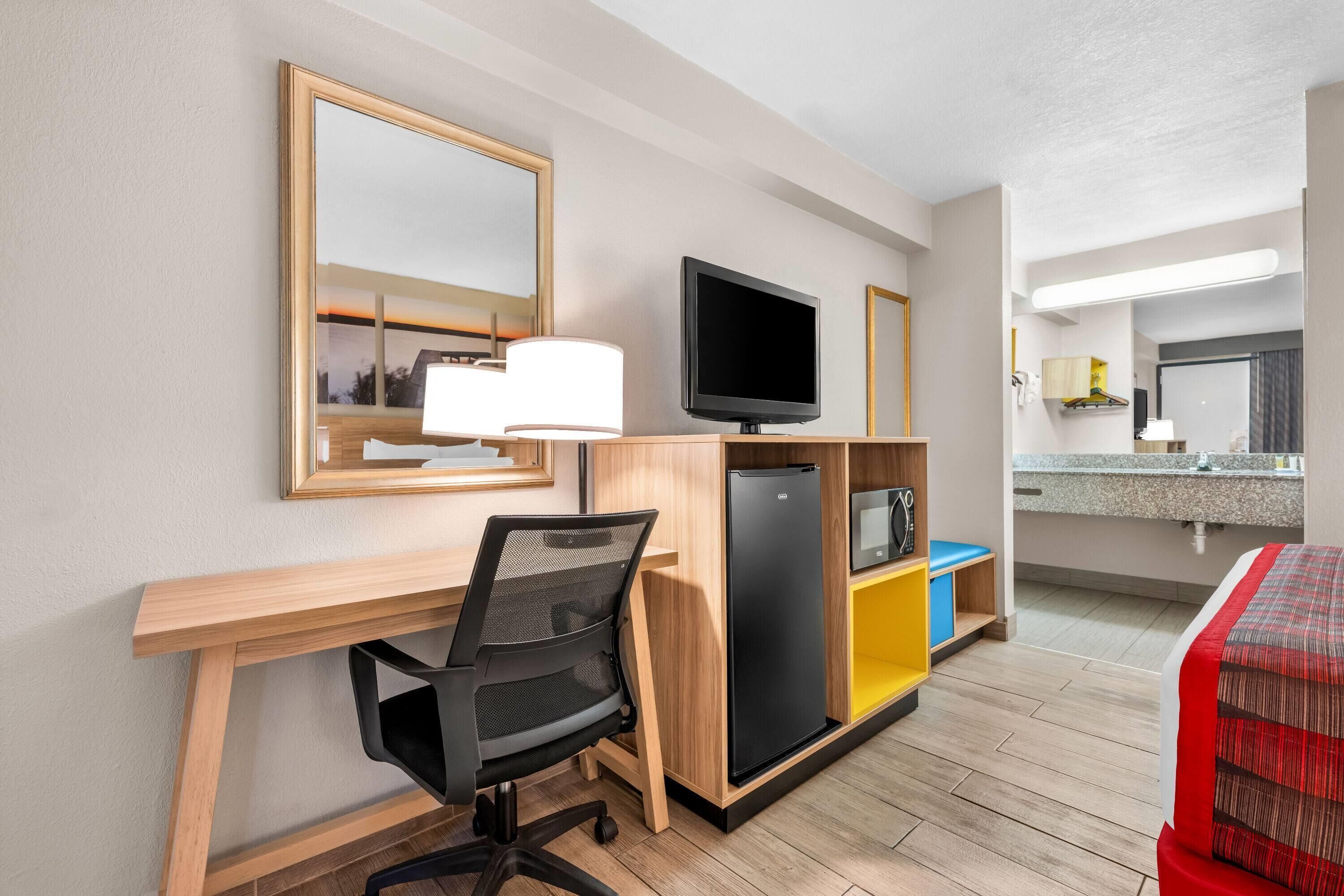 A hotel room with a wooden desk, a black office chair, a television, a mini-fridge, and a bed with a red and white striped blanket.