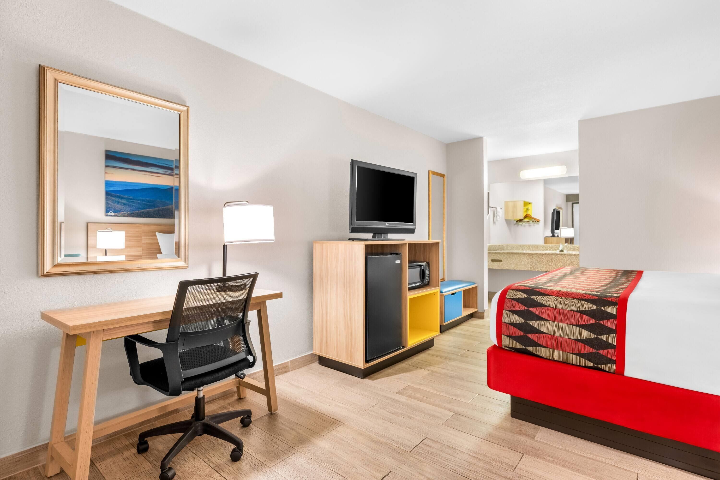Camera d'albergo con una scrivania in legno, una sedia da ufficio nera, una televisione su un mobiletto in legno, un letto con coperta a motivi rossi e beige e un angolo cottura con forno a microonde e frigorifero.