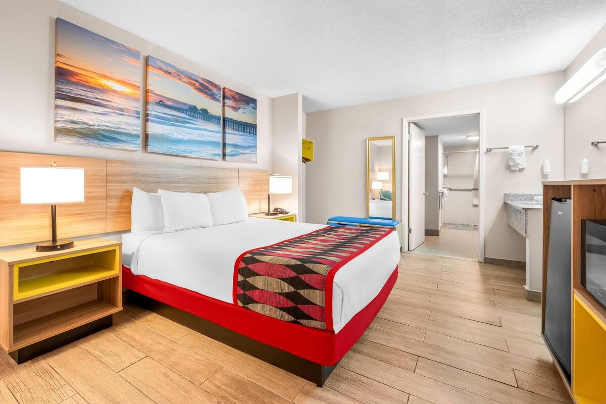 A hotel room with a bed, bedside tables, a mirror, a TV, and artwork on the wall.