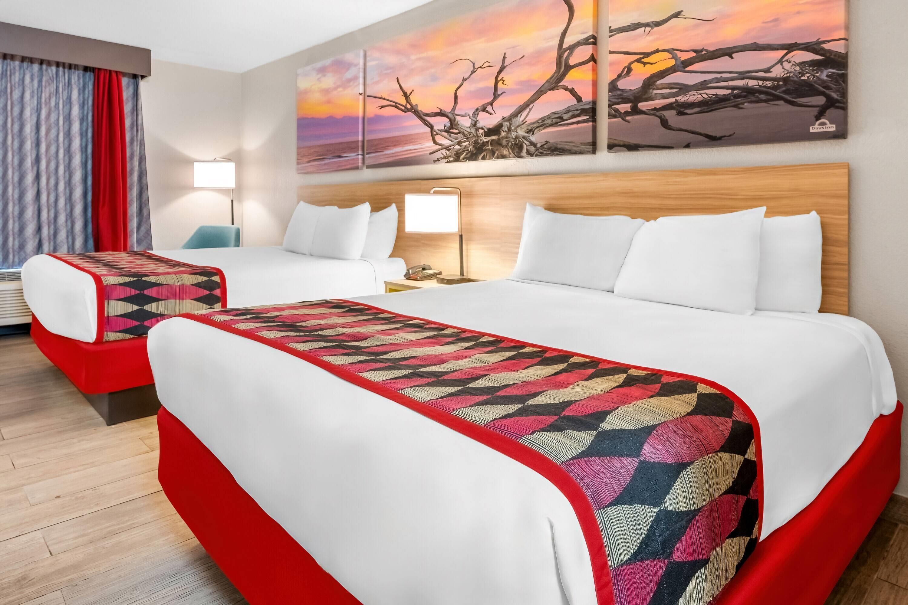 Habitación de hotel con dos camas, decoración en tonos rojo y blanco, un ventanal con cortinas y un cuadro de atardecer en la pared.
