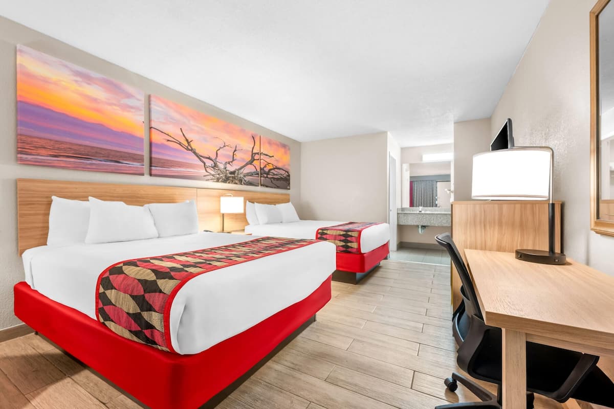 A hotel room with a large bed, a desk, and a chair. There are two paintings on the wall and a mirror.