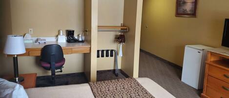 A hotel room with a bed, desk, chair, lamp, coffee maker, and a small refrigerator.