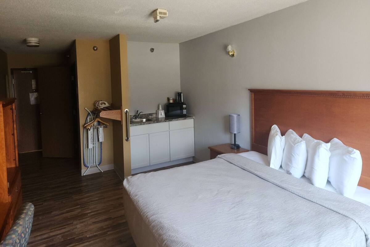 Ein Hotelzimmer mit einem großen Bett, einem Holzkopfende, einem kleinen Schreibtisch mit Lampe, einer Küchenzeile und einem durchgehenden Türspalt sichtbaren Badezimmer.