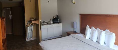 A hotel room with a large bed, a wooden headboard, a small desk with a lamp, a kitchenette, and a bathroom visible through an open door.