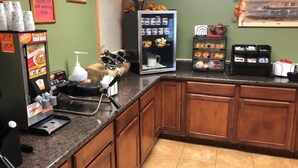 Una zona de cocina con una plancha para gofres, cafetera y un refrigerador lleno de bebidas.