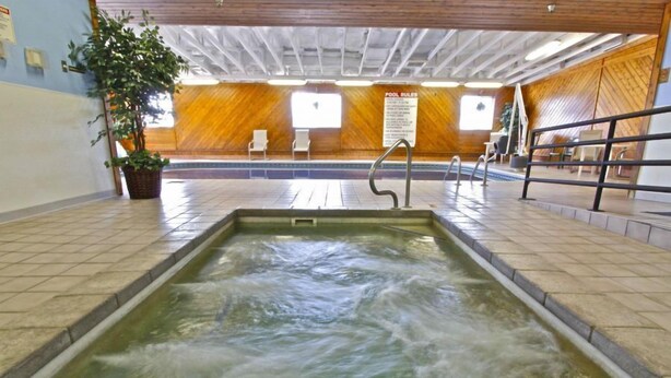 Una piscina interior con techo de madera y suelo de baldosas, rodeada de sillas y un cartel con las normas de la piscina.