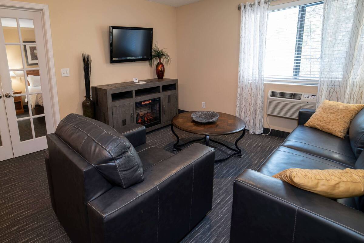 A modern living room with a leather sofa, a round coffee table, a flat-screen TV mounted on the wall, and a fireplace.