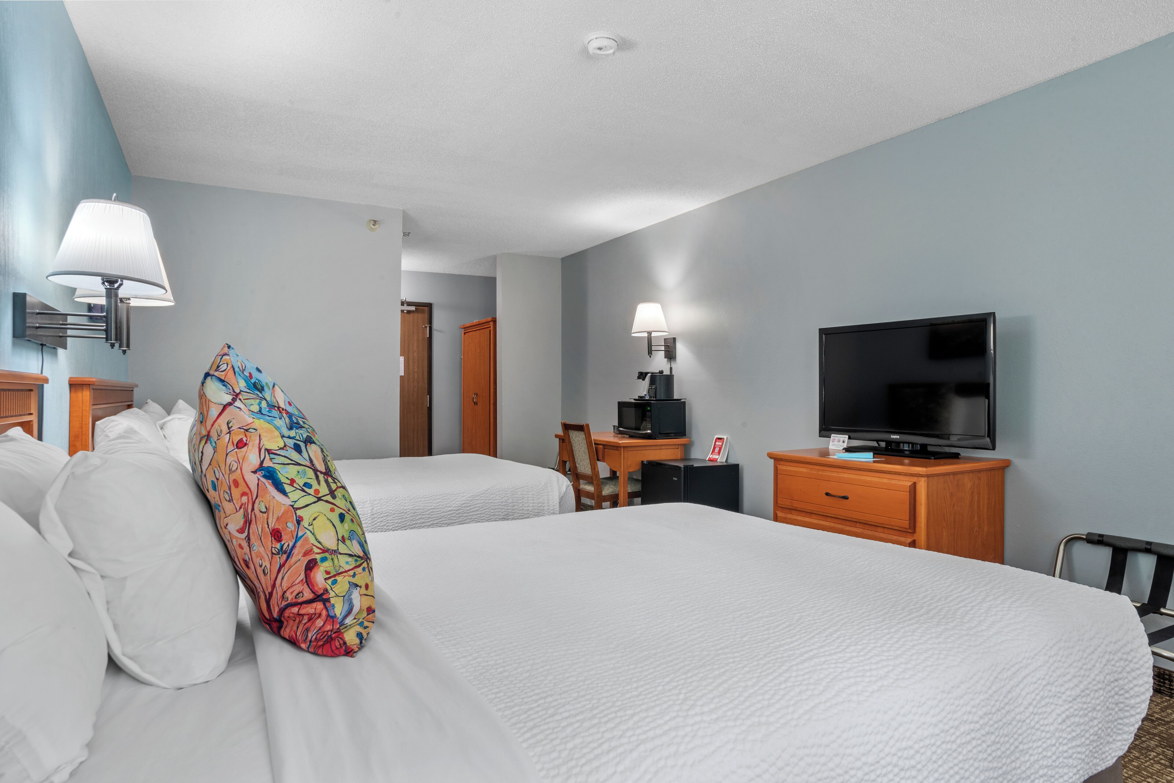 A hotel room with two beds, a TV, a desk with a chair, and a lamp.