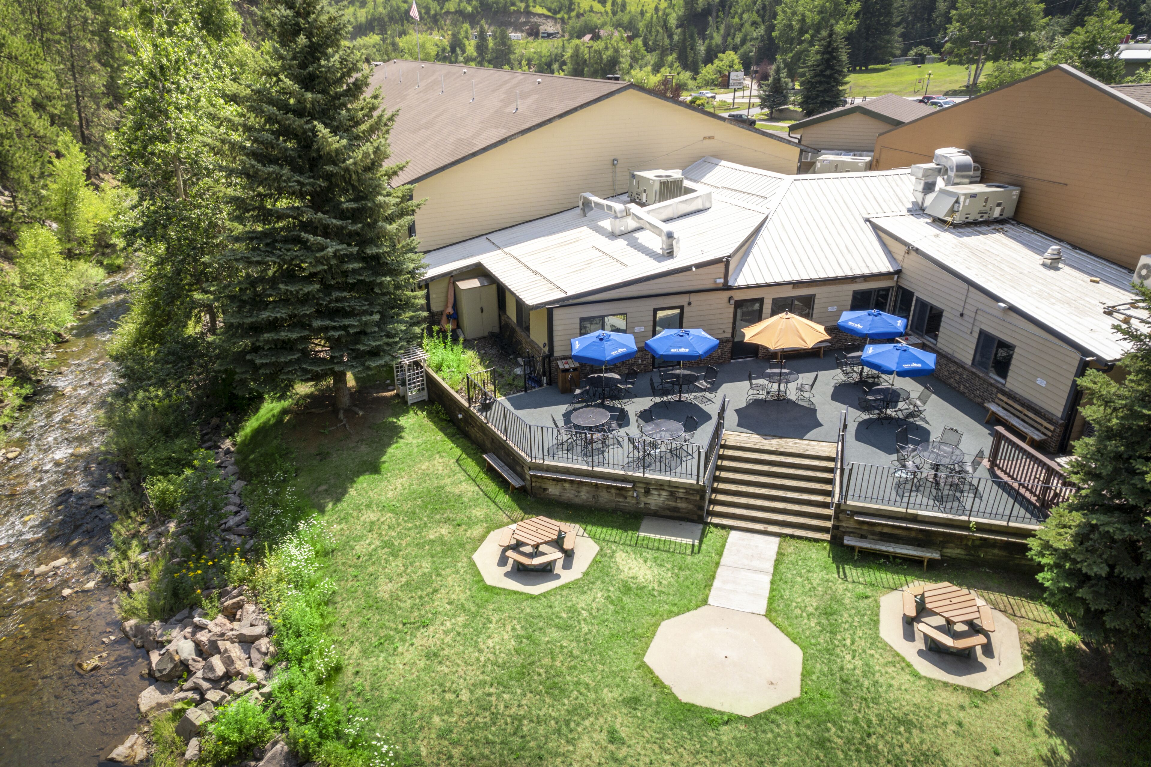 An outdoor patio area with tables and umbrellas, surrounded by a grassy lawn and a river.