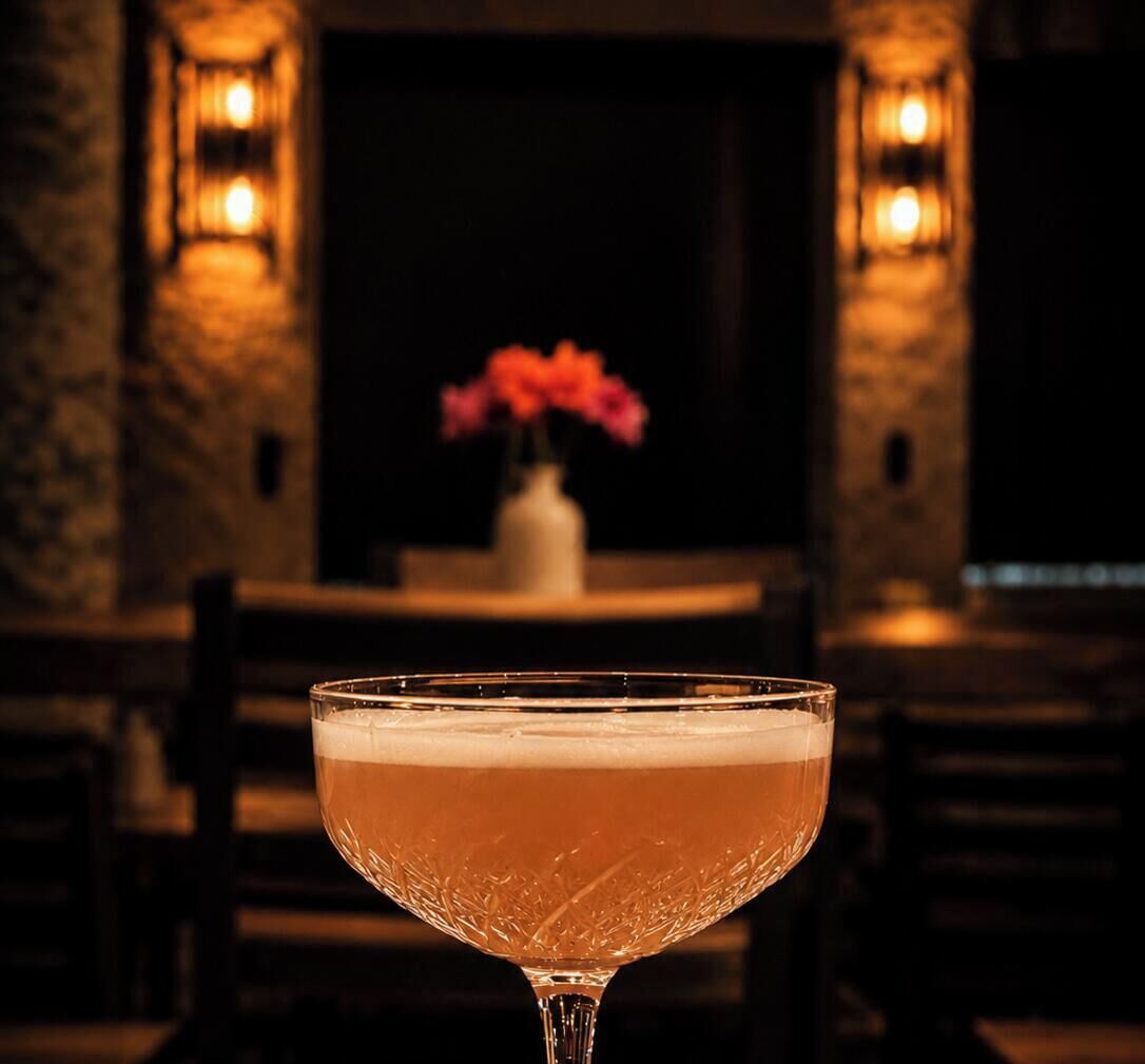 Un cocktail servi dans un verre dont le bord est texturé, présenté dans un décor aux murs de pierre et à la lumière tamisée, avec un vase de fleurs en arrière-plan.
