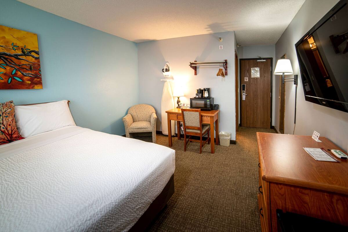 A hotel room with a bed, a desk with a lamp, a chair, a TV, and a painting on the wall.