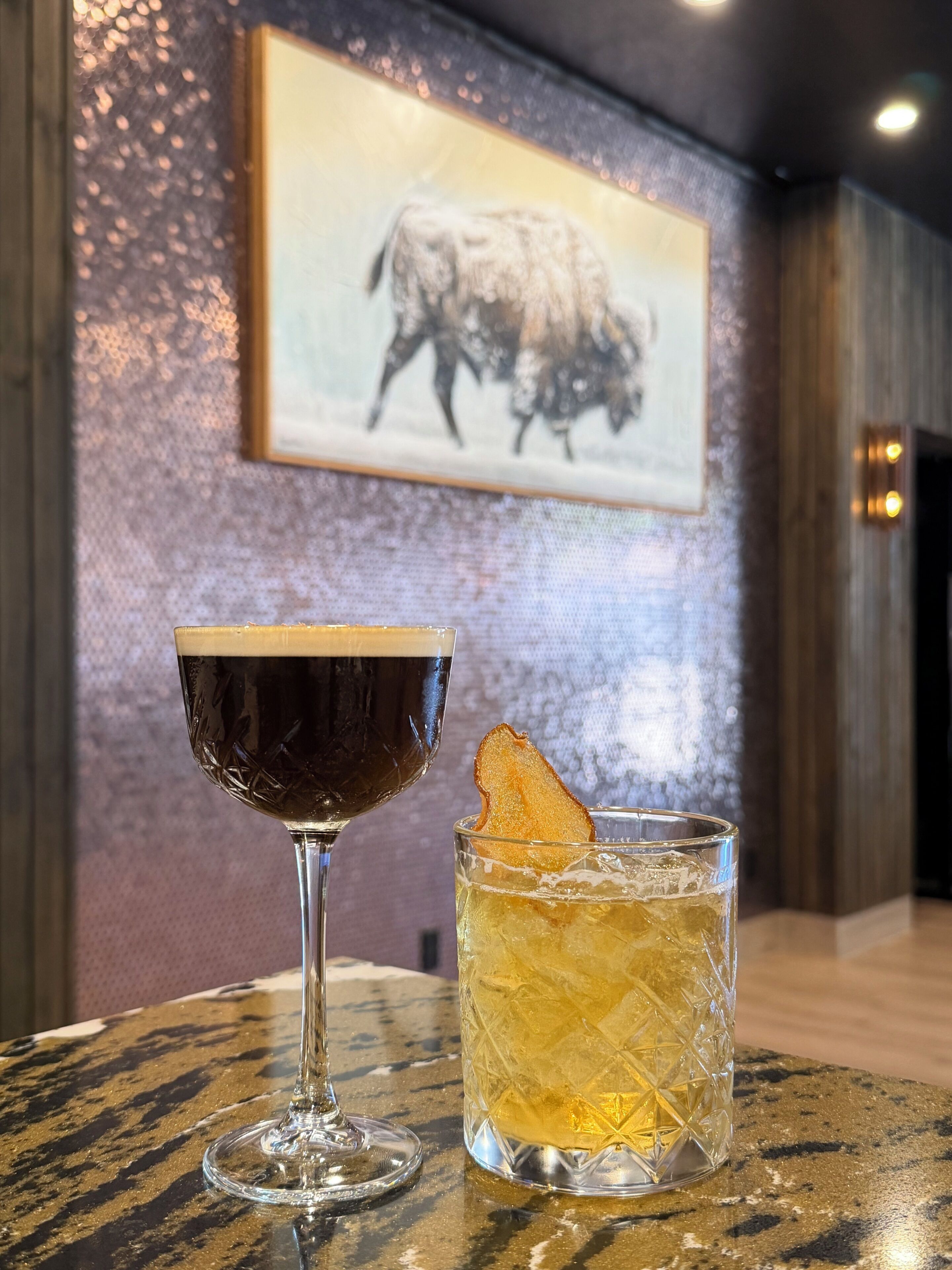 Two glasses of beverages on a table with a bull painting in the background.