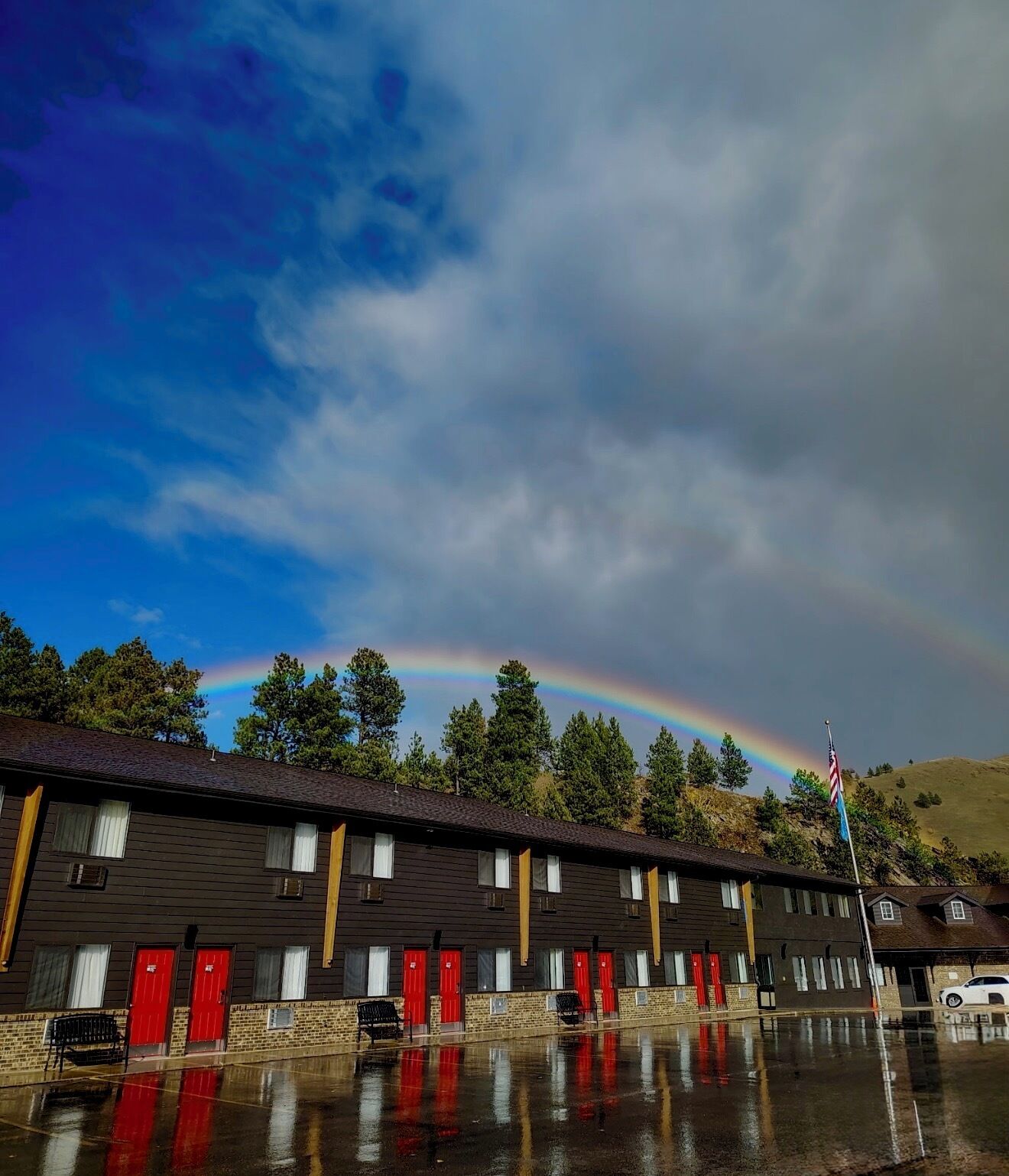 A row of motel rooms with red doors, a flagpole with an American flag, and a rainbow in the background.