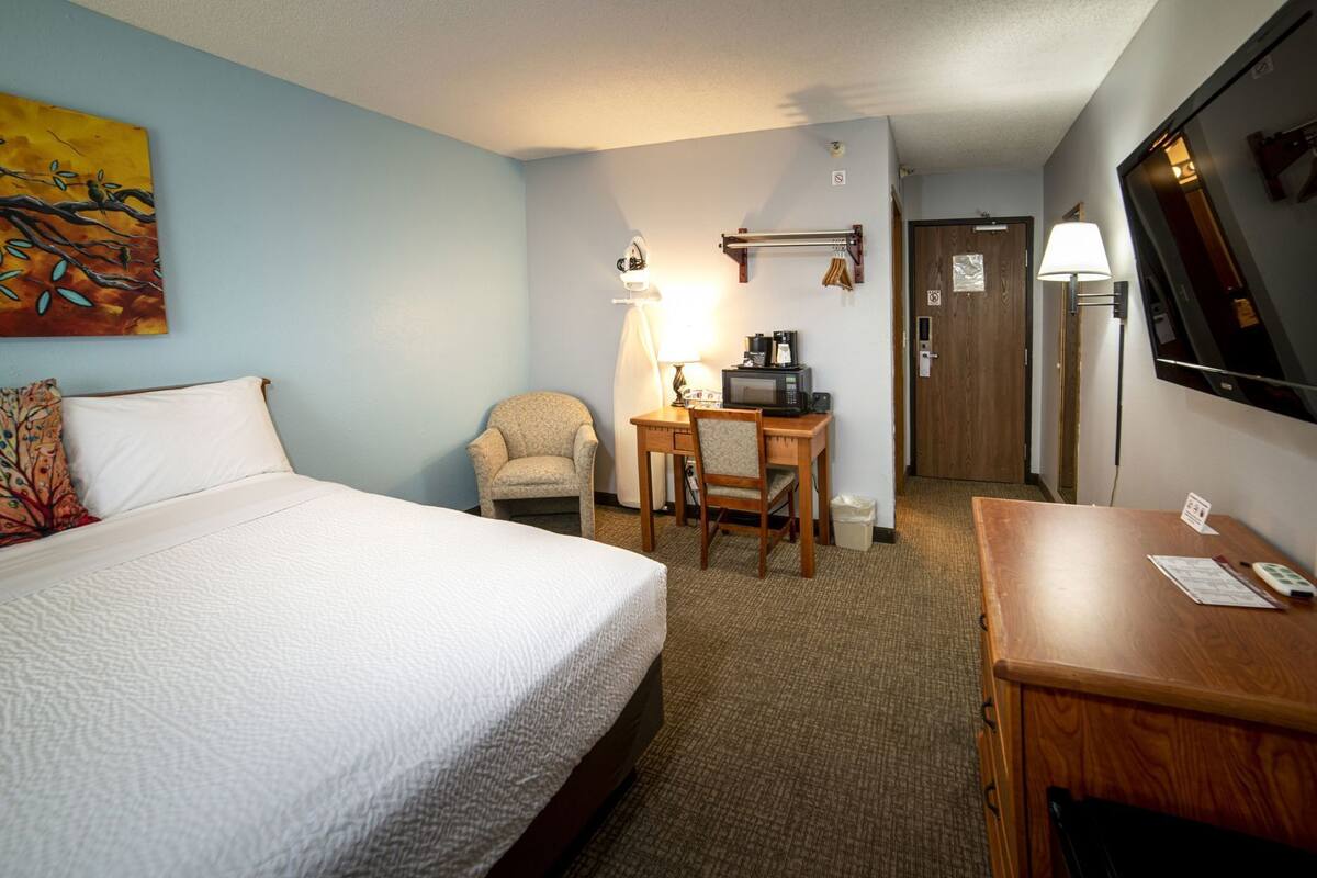 A hotel room with a bed, a desk with a chair, a TV, and a painting on the wall.