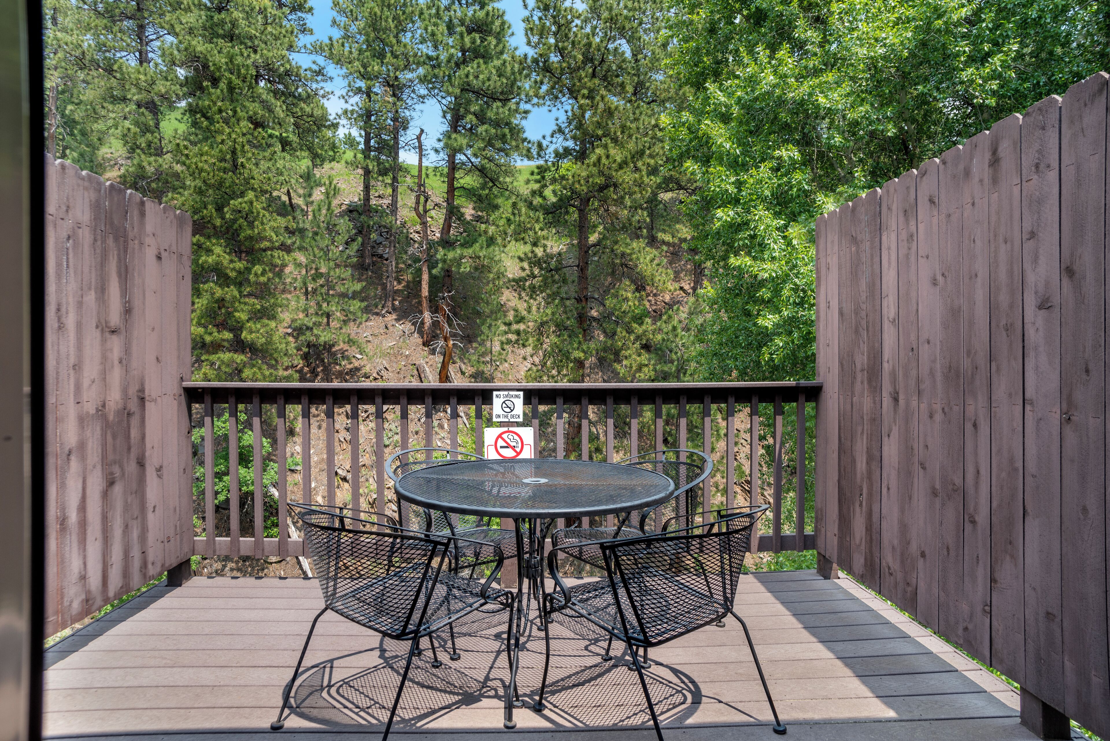 A wooden deck with a round metal table and four chairs, surrounded by a wooden fence and trees.