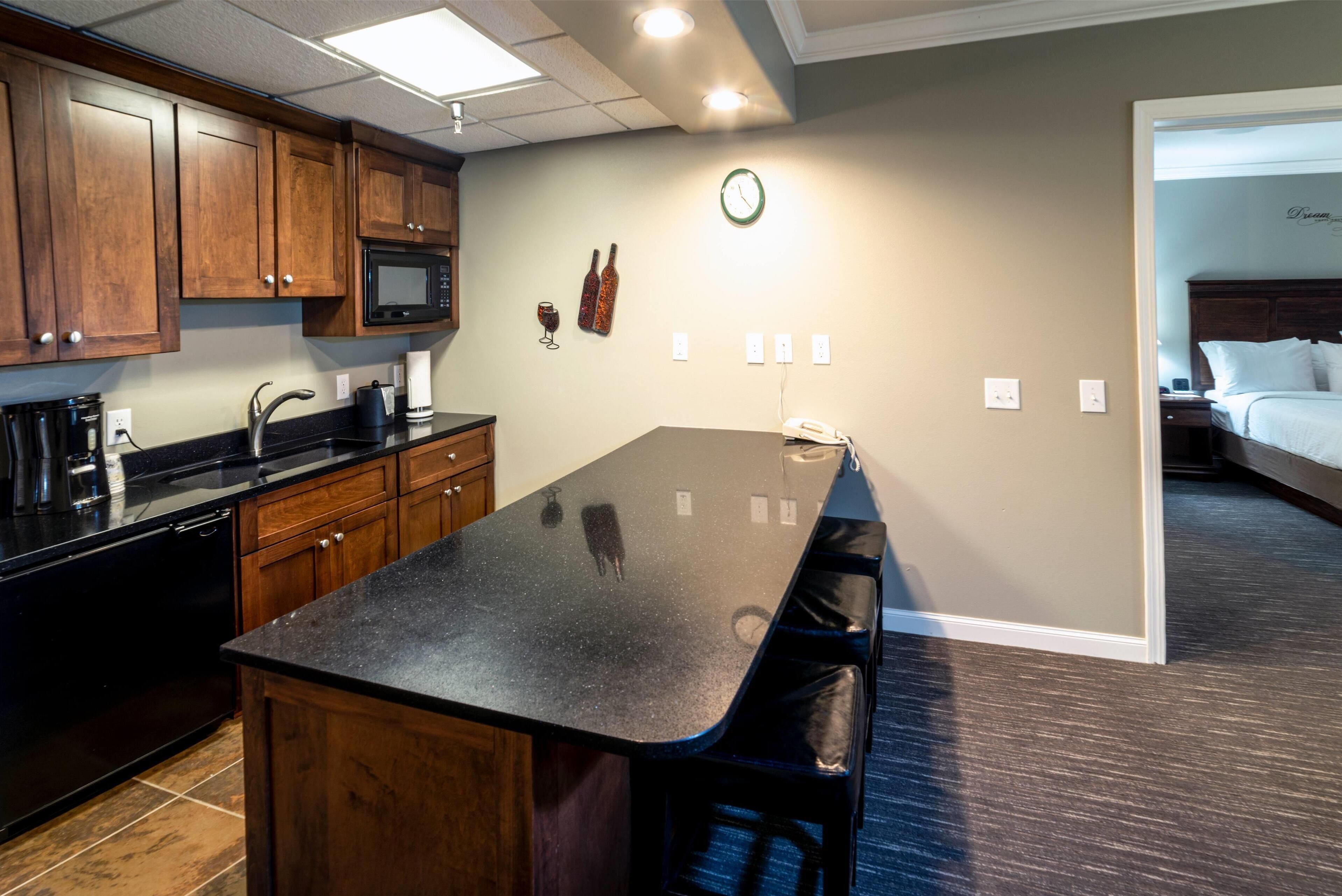 A kitchen with wooden cabinets, a black countertop, and a microwave. There is a clock on the wall and a bed visible in the adjacent room.