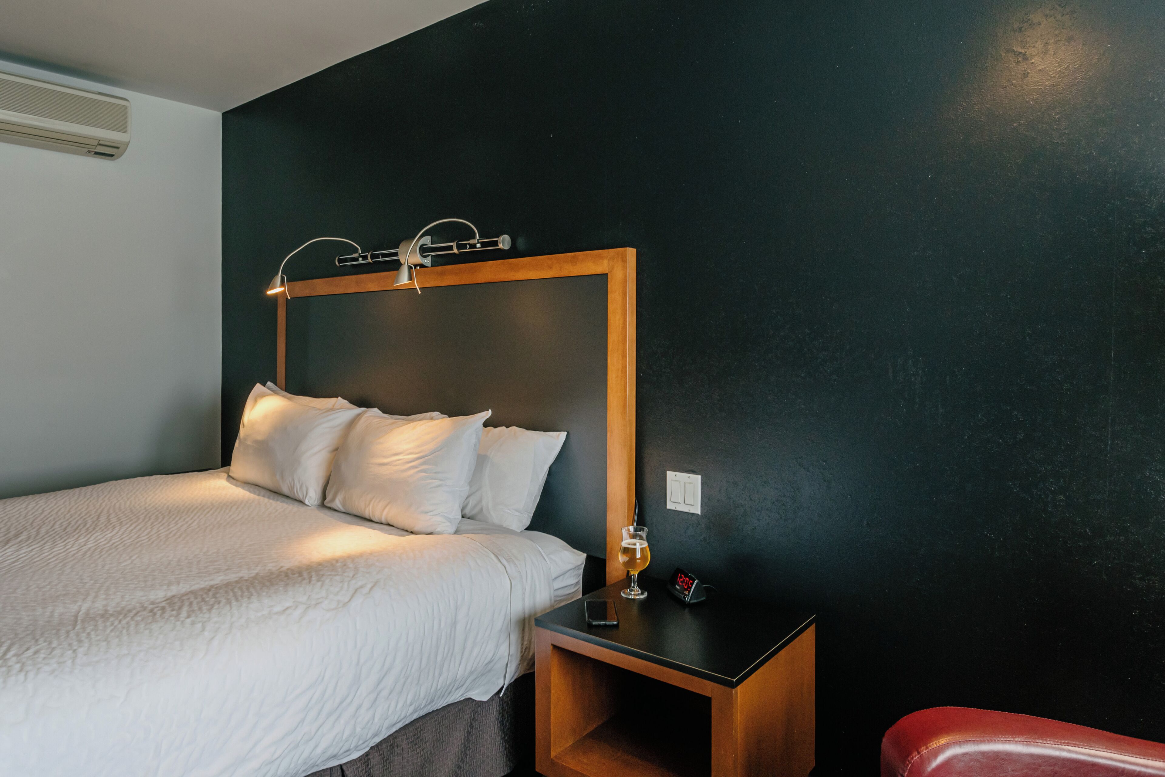A neatly made bed with white linens, a bedside table with a glass of drink and a clock, and a red chair.