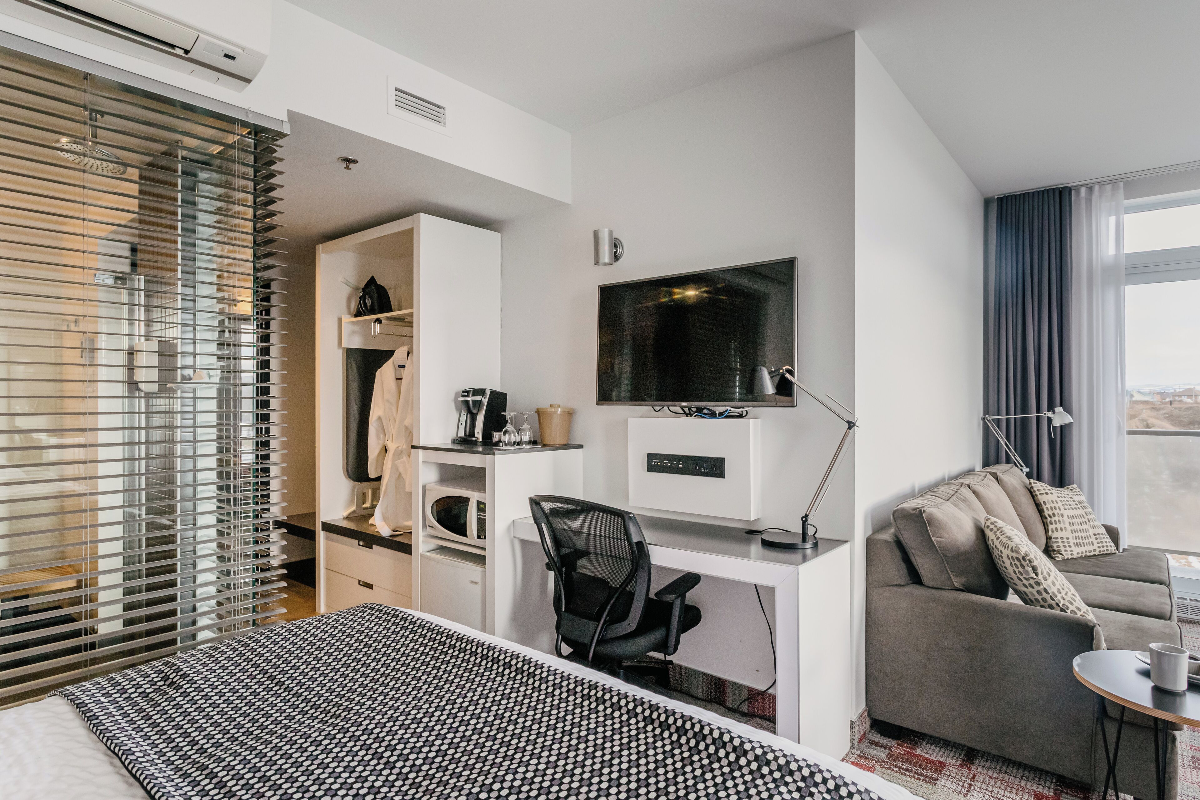 Een moderne hotelkamer met een bed, een bureau met een computer, een televisie, een bank en een raam met zonwering.