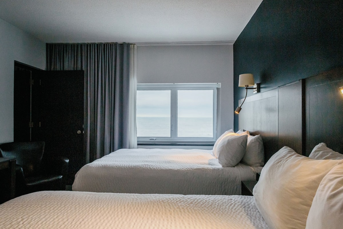 Habitación de hotel con dos camas, un escritorio y vista al mar a través de un ventanal grande.