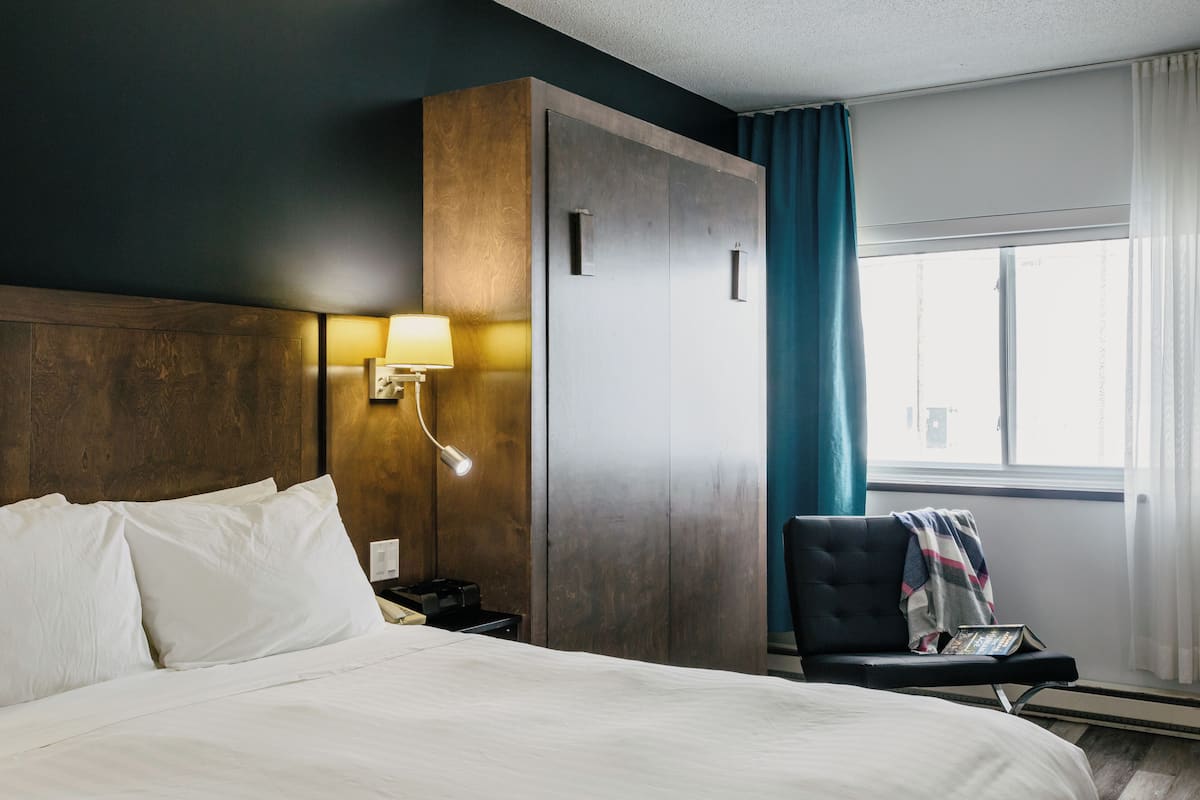 Una habitación de hotel con cama, mesita de noche, lámpara, armario, silla y ventana con cortinas.