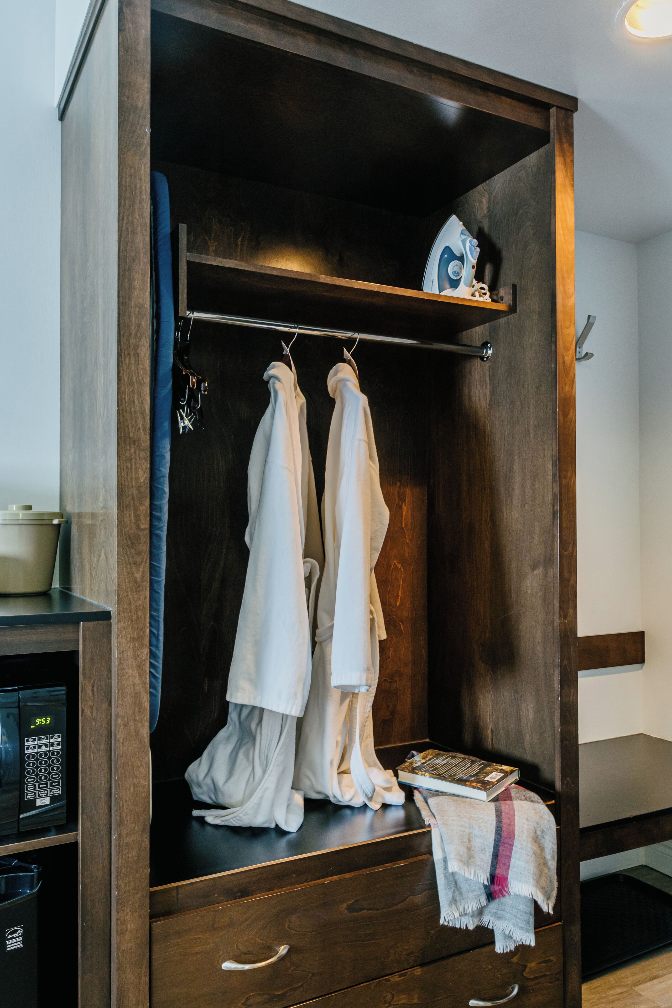A wooden wardrobe with a hanging rack, a shelf with an iron, and a microwave on a counter.