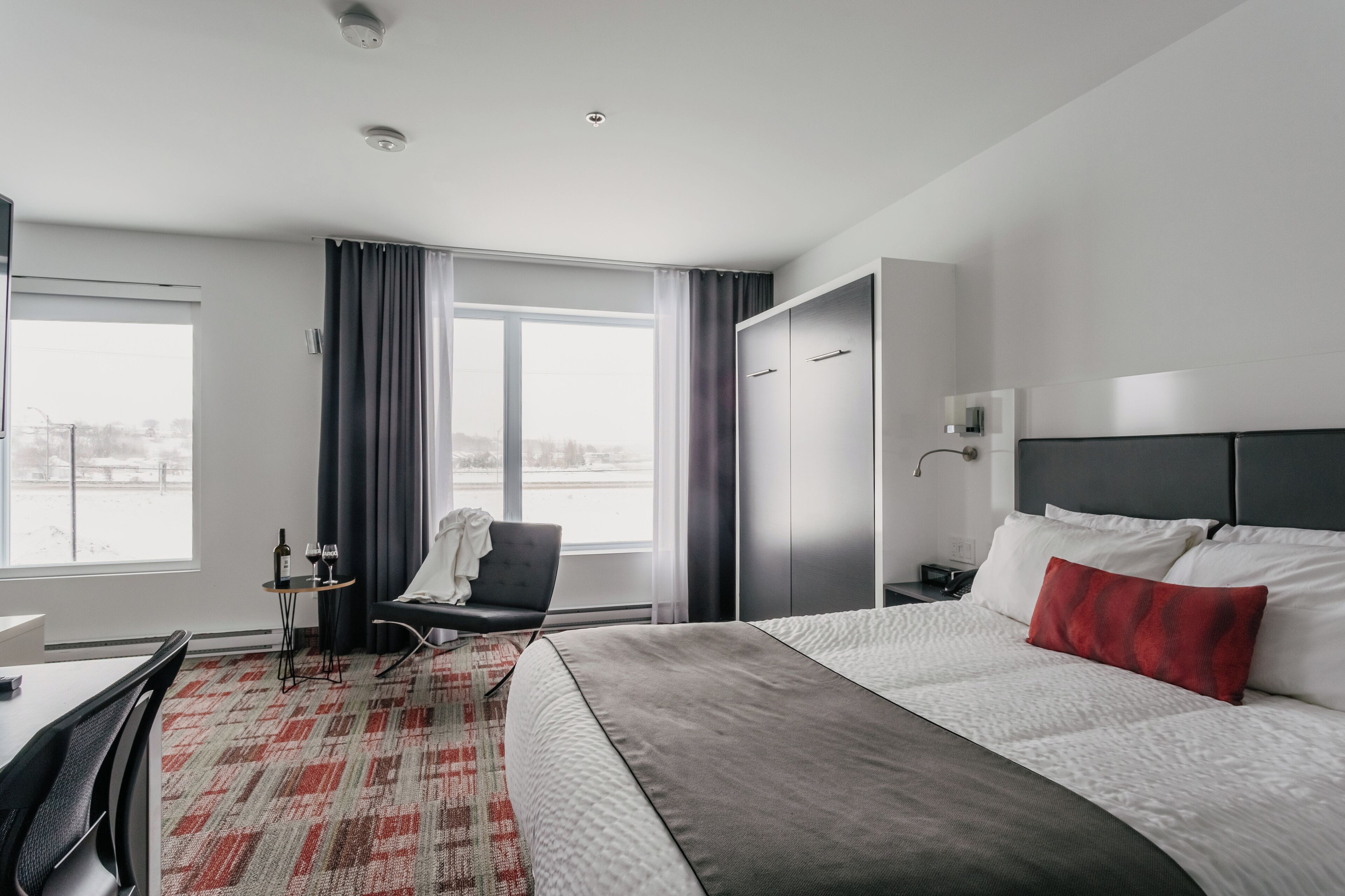 Habitación de hotel con una cama grande, un escritorio con una silla, una mesita con una botella y vasos, otra silla, un armario y una ventana con cortinas.