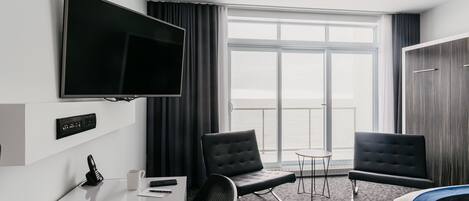 A hotel room with a flat-screen TV, a desk with a phone, two chairs, a small table, and a large window with a view.