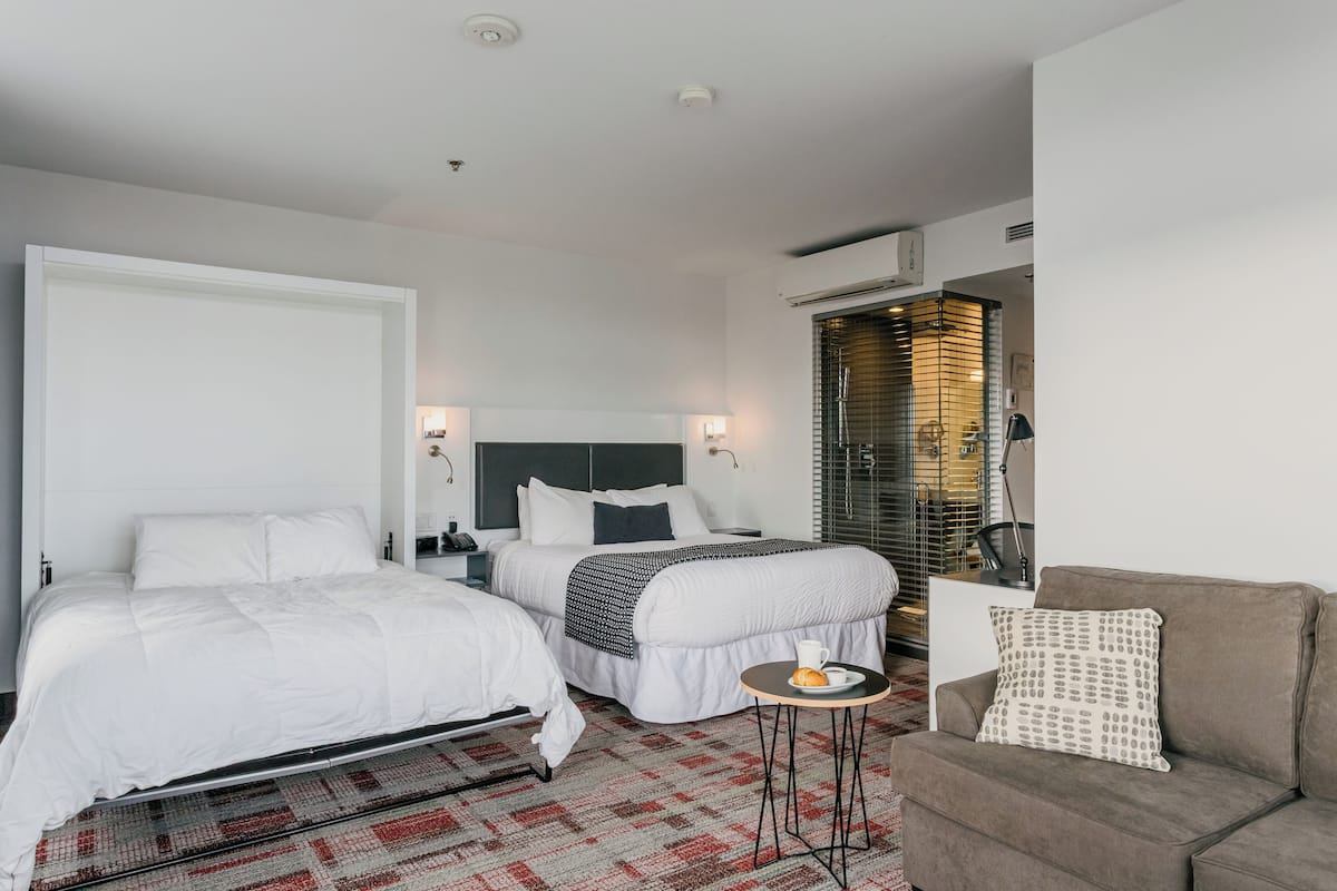 Una habitación de hotel moderna con dos camas, un sofá, una mesita con una taza y un pastel, y un ventanal grande con persianas.