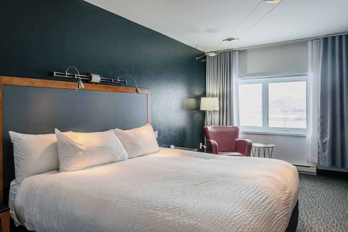 Habitación de hotel con una cama grande, una silla roja, una mesita y vistas al exterior a través de una ventana.