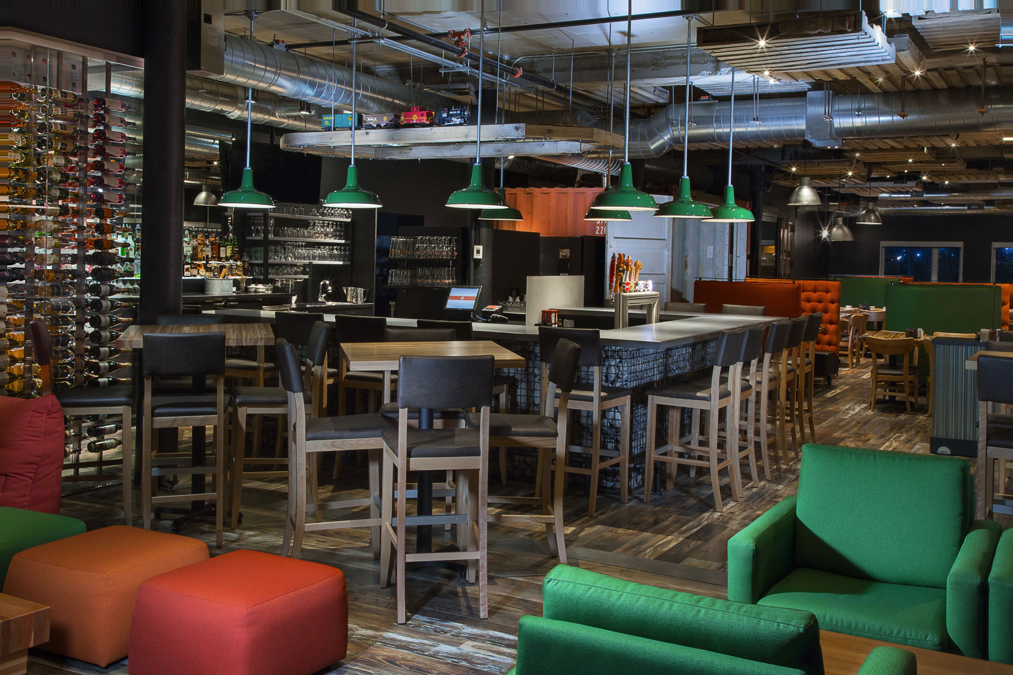 Un bar moderne avec des meubles en bois, des sièges verts et orange, et une variété de bouteilles et de verres exposés sur des étagères.