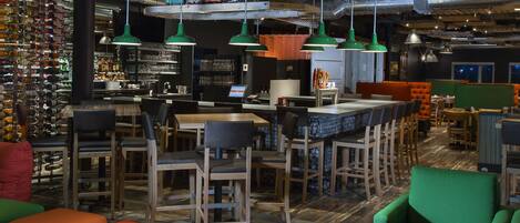 Un bar moderne avec des meubles en bois, des sièges verts et orange, et une variété de bouteilles et de verres exposés sur des étagères.