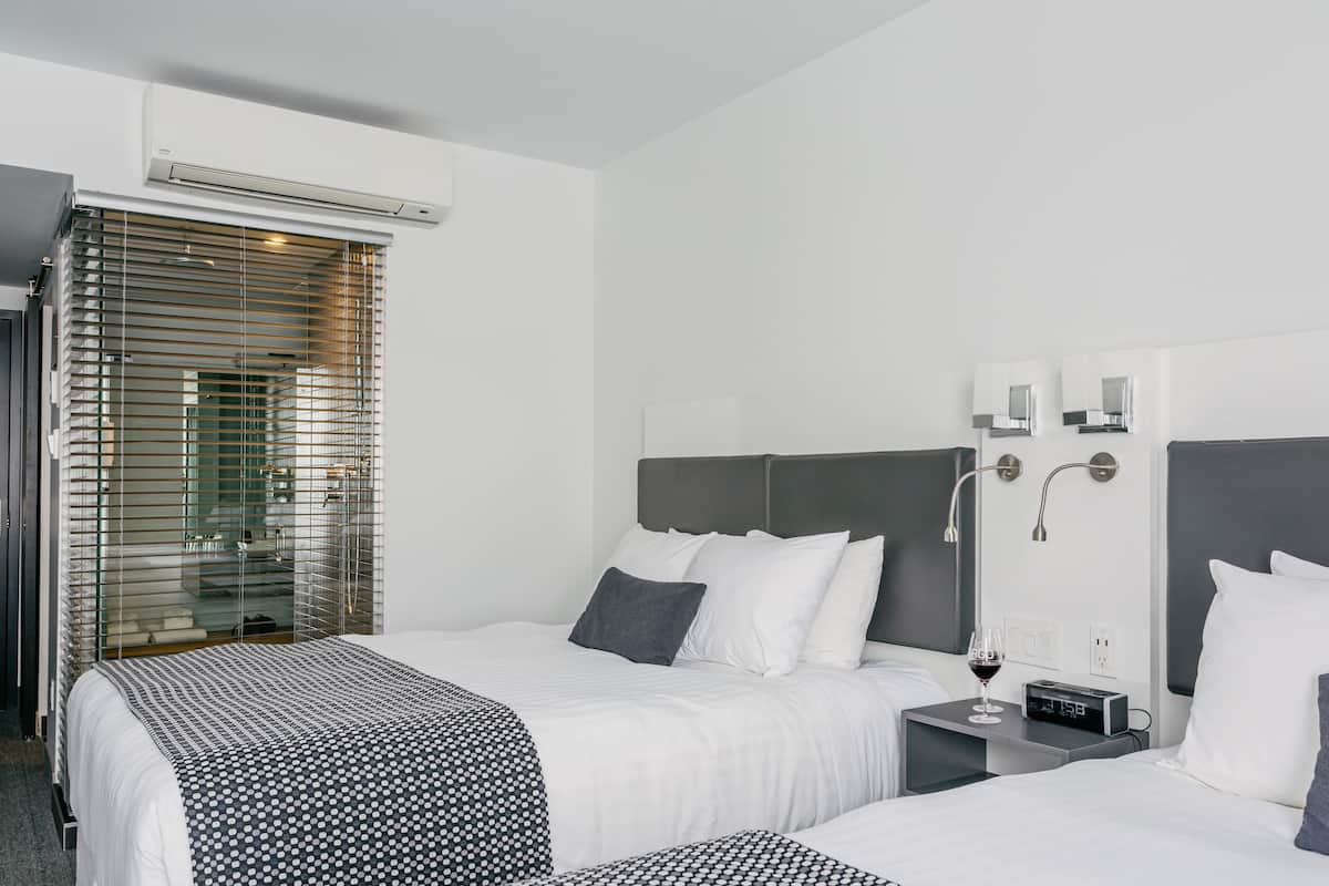 Una habitación de hotel moderna con dos camas, una mesita de noche con una copa de vino, un reloj digital y un ventanal grande con persianas.